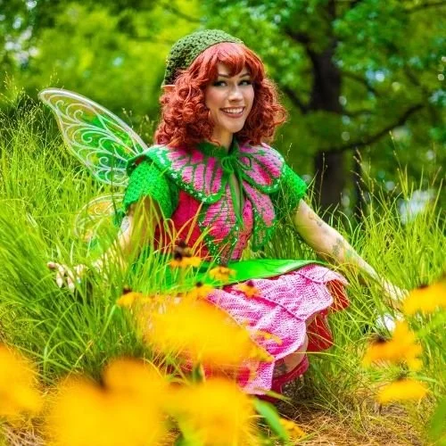 A woman dressed as a fairy with pink curly hair, green wings, and a green hat sitting in a grassy field with yellow flowers, smiling.