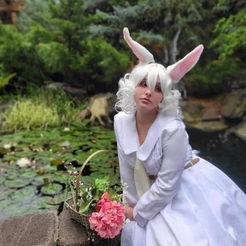 Person dressed as a white rabbit with long ears and curly white hair, holding pink flowers, outdoors by a pond with lily pads and green trees in the background.