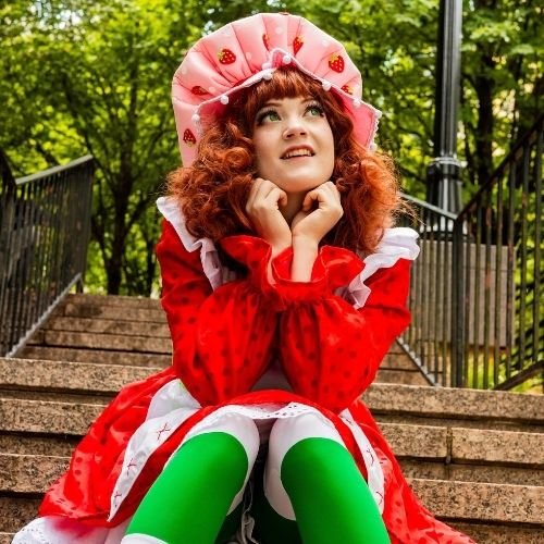 A woman dressed as Strawberry Shortcake sitting on outdoor steps, looking up with a dreamy expression, surrounded by green trees.