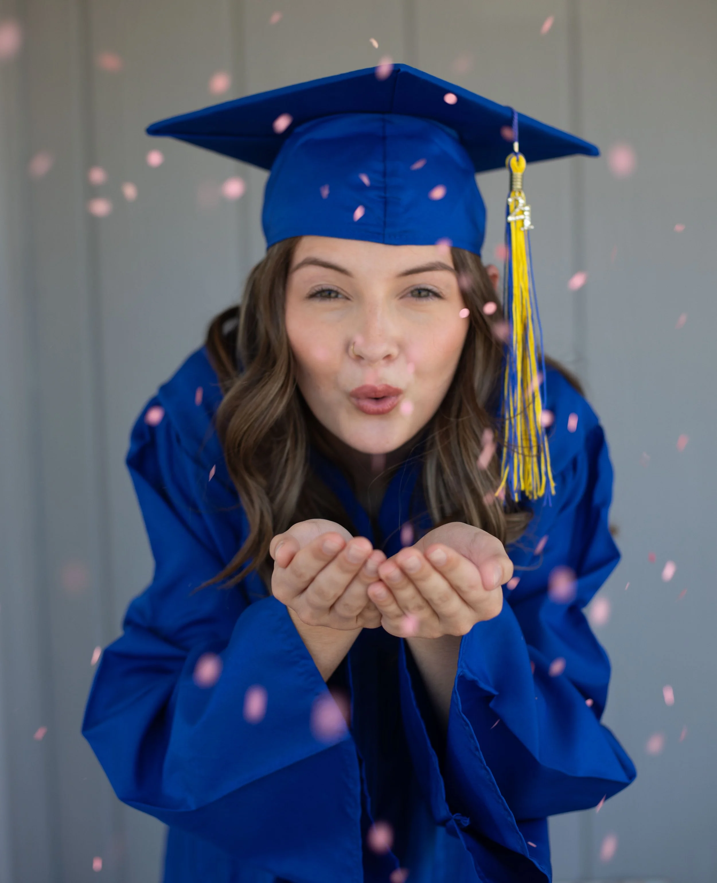 Phelan, CA senior photo of a girl with blue cap and gown blowing pink confetti to celebrate her graduation in the High desert, by family photographer.