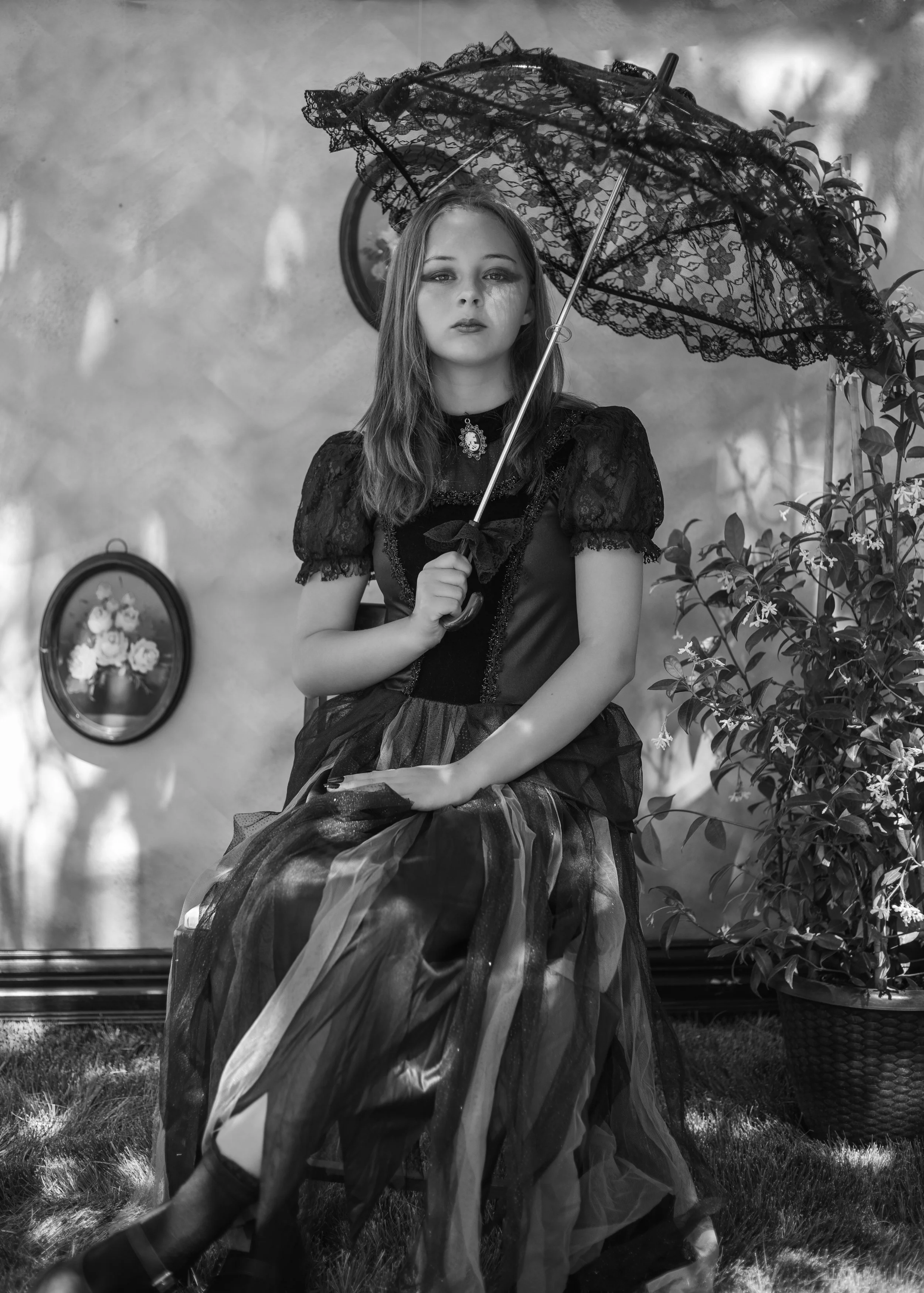 Girl cosplay victorian clothing holding umbrella, sitting surrounded by plants and old photos on a wall. Victorville photographer for cosplay and whimsy, fantasy portraits.