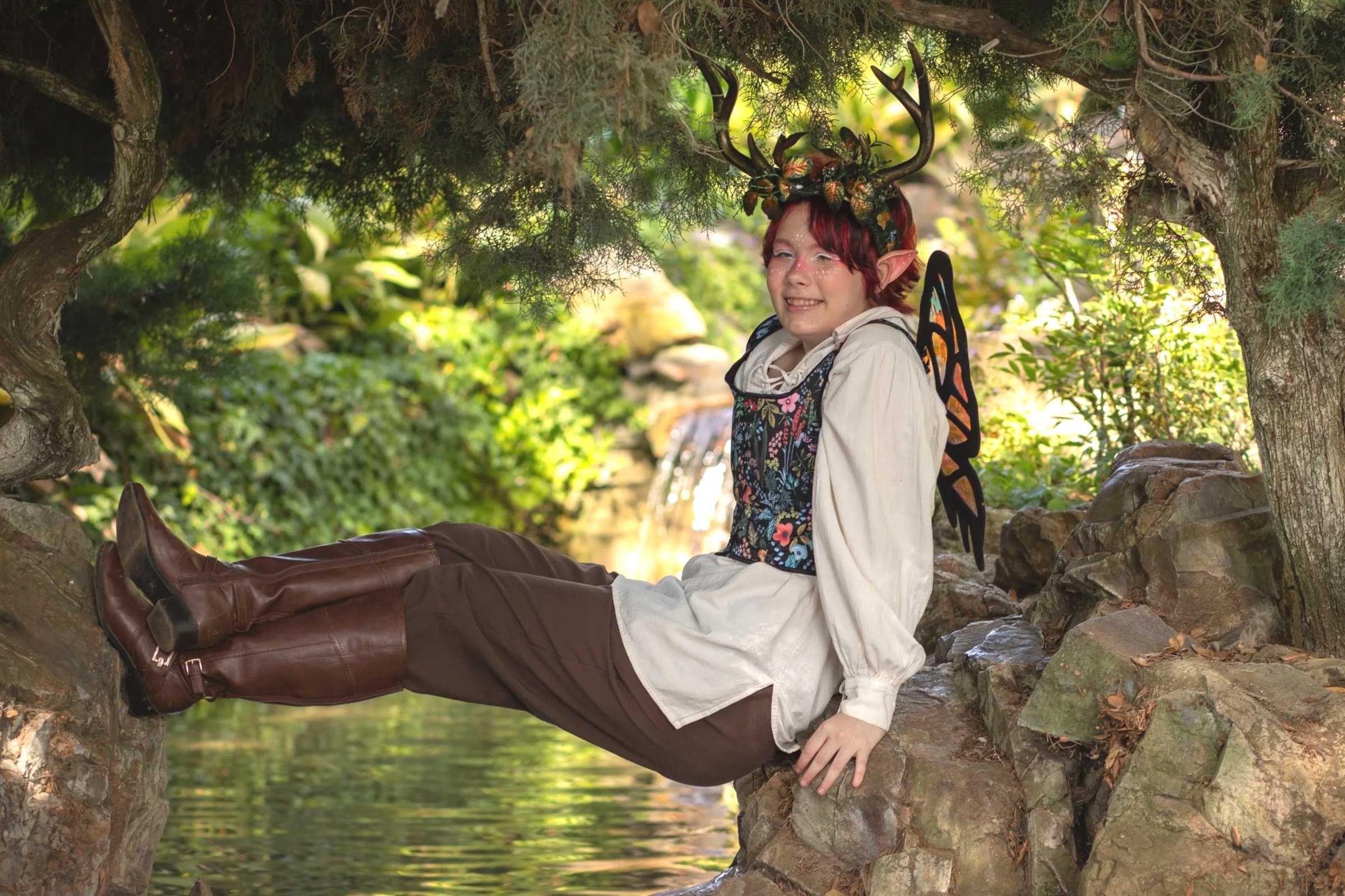 A person dressed as a fantasy elf with deer antlers, black wings, and red hair, sitting on a tree branch in a garden with lush green foliage. Fantasy photographer in high desert and inland empire, CA.