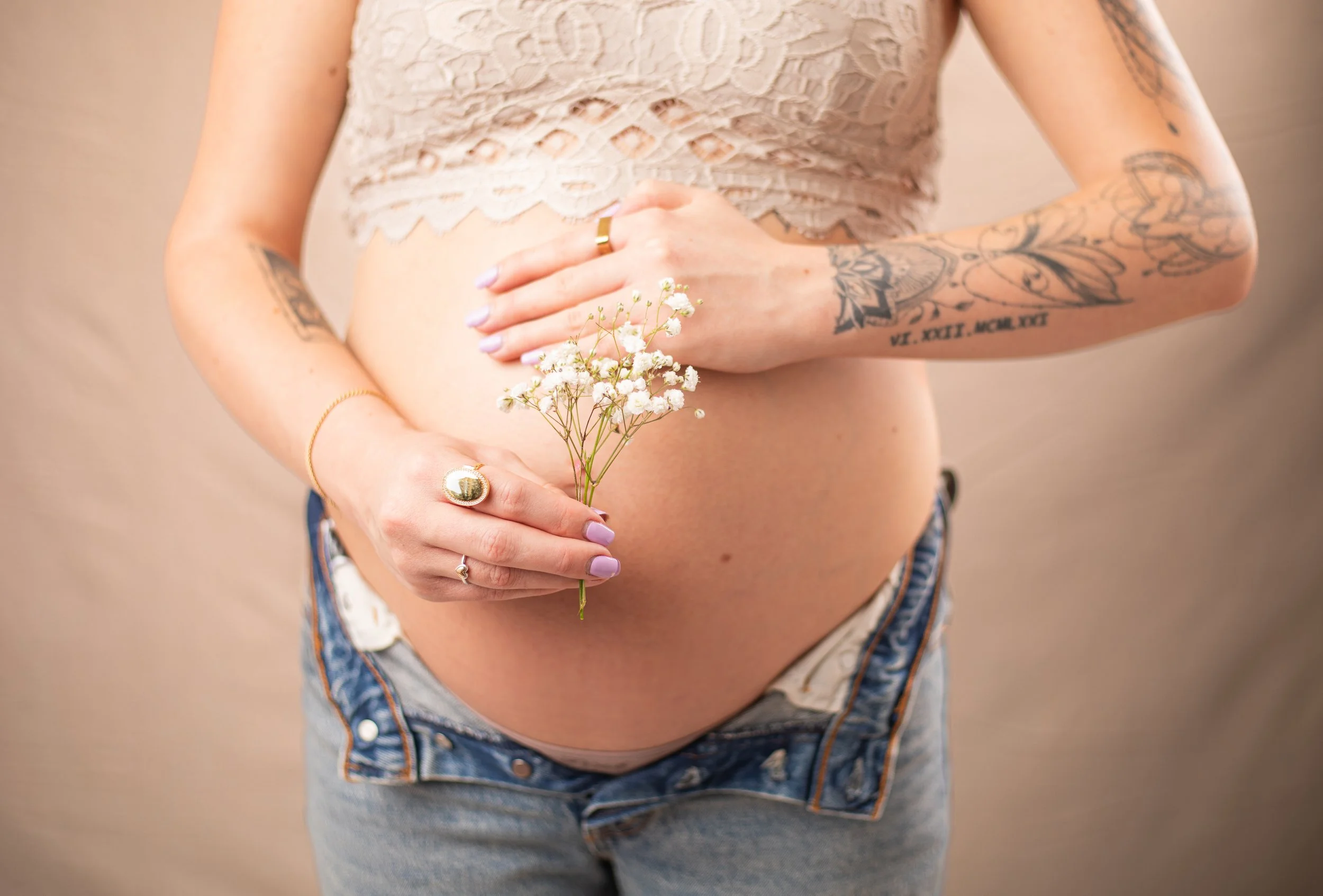 High desert photography of pregnant woman holding her belly and wearing jeans with a lacy top while holding flowers and wearing rings.