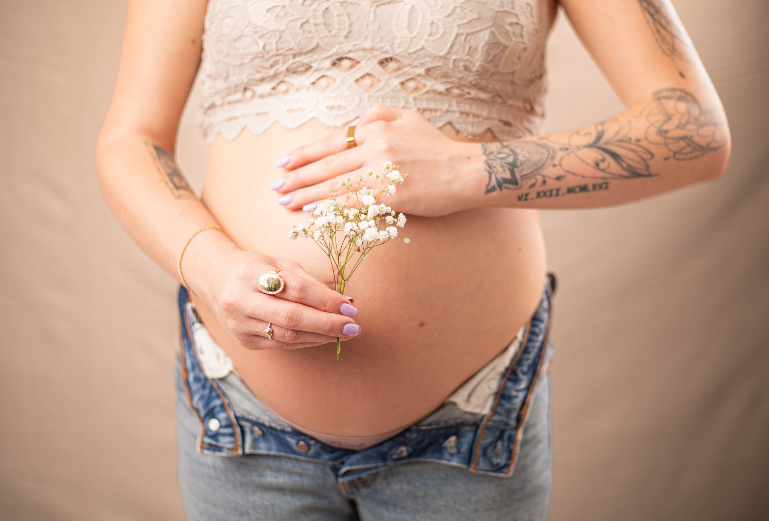 pregnany woman holding her belly and wearing jeans and a lacy top whilte holding flowers and wearing rings. phelan family photographer.