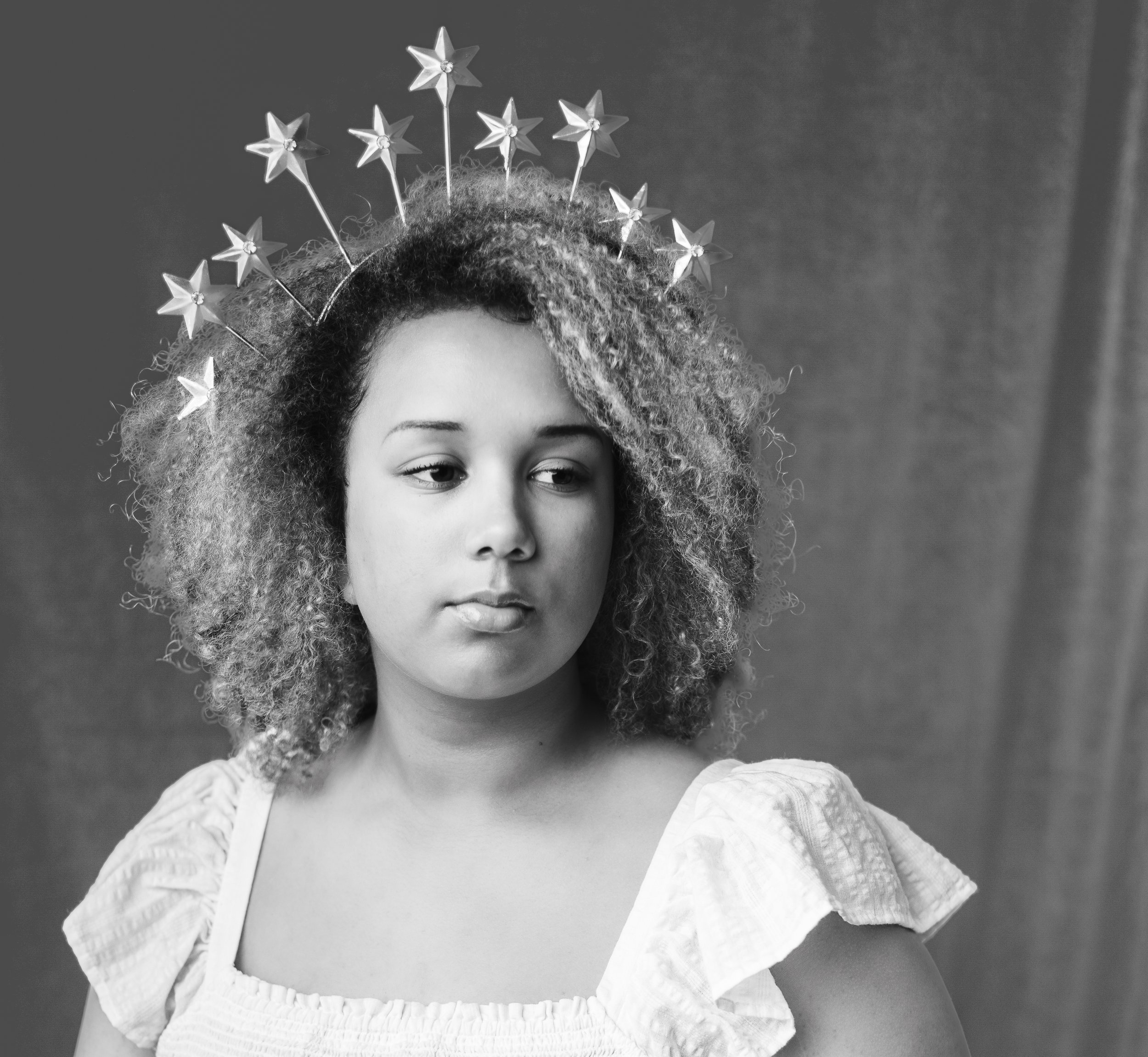 Black girl sitting and wearing her nautral hair with a star crown and white dress, looking to the side with boredom in her eyes. Photographer for families in phelan, Ca