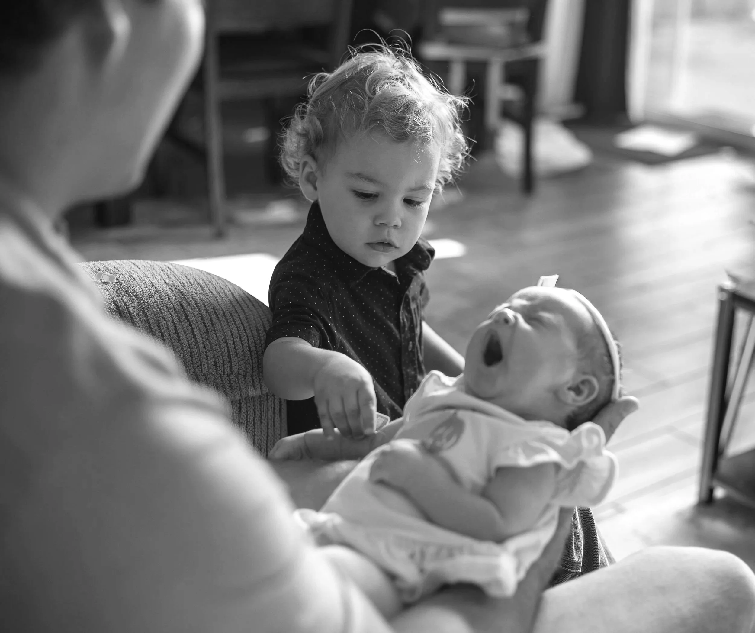 Toddler boy touching newborn baby sitter while baby yawns. In home lifestyle session by high desert family photographer.
