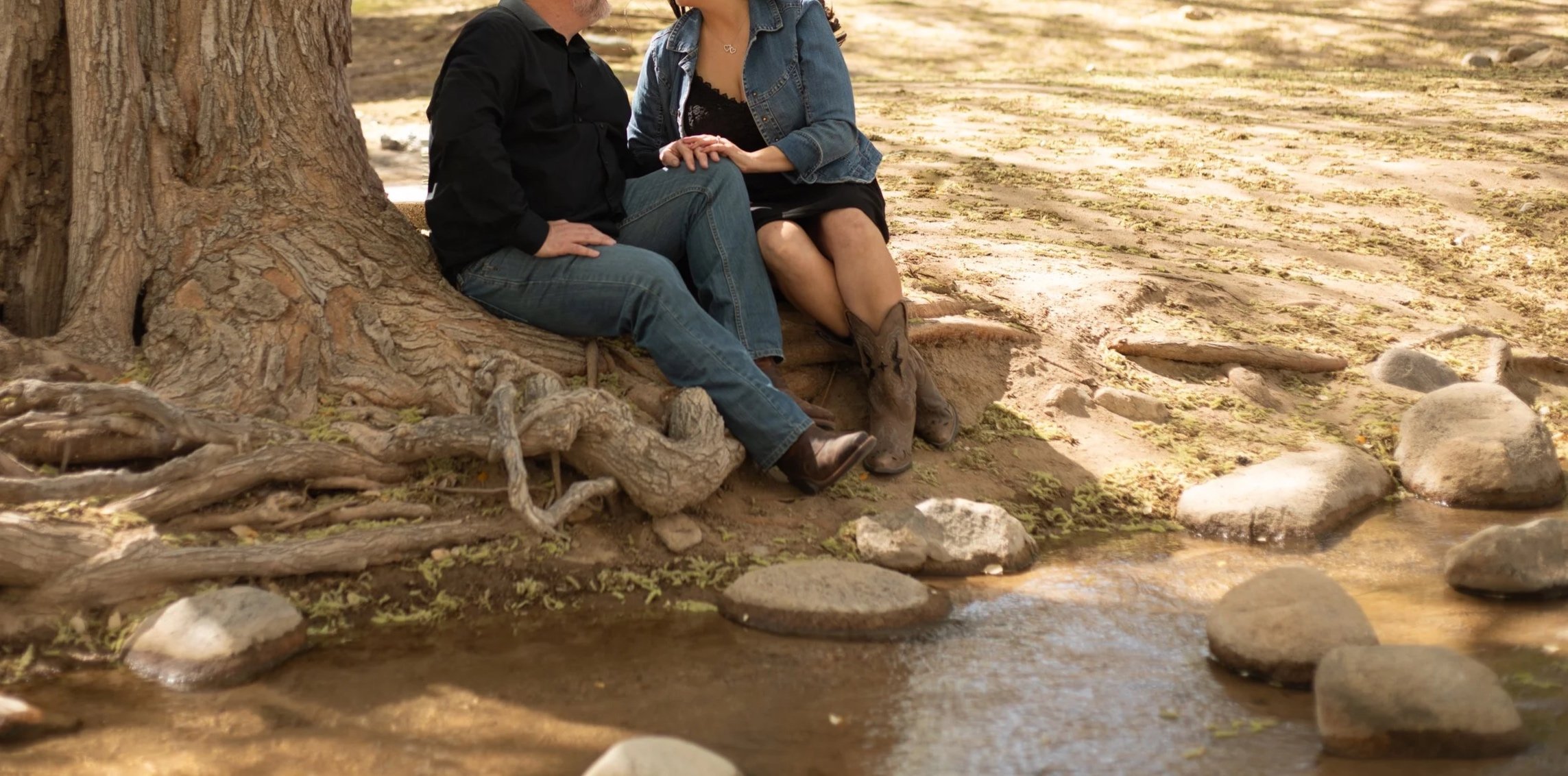 Two people sitting on a tree root by a creek, holding hands, with a donkey nearby. Hesperia Photographer who loves people.