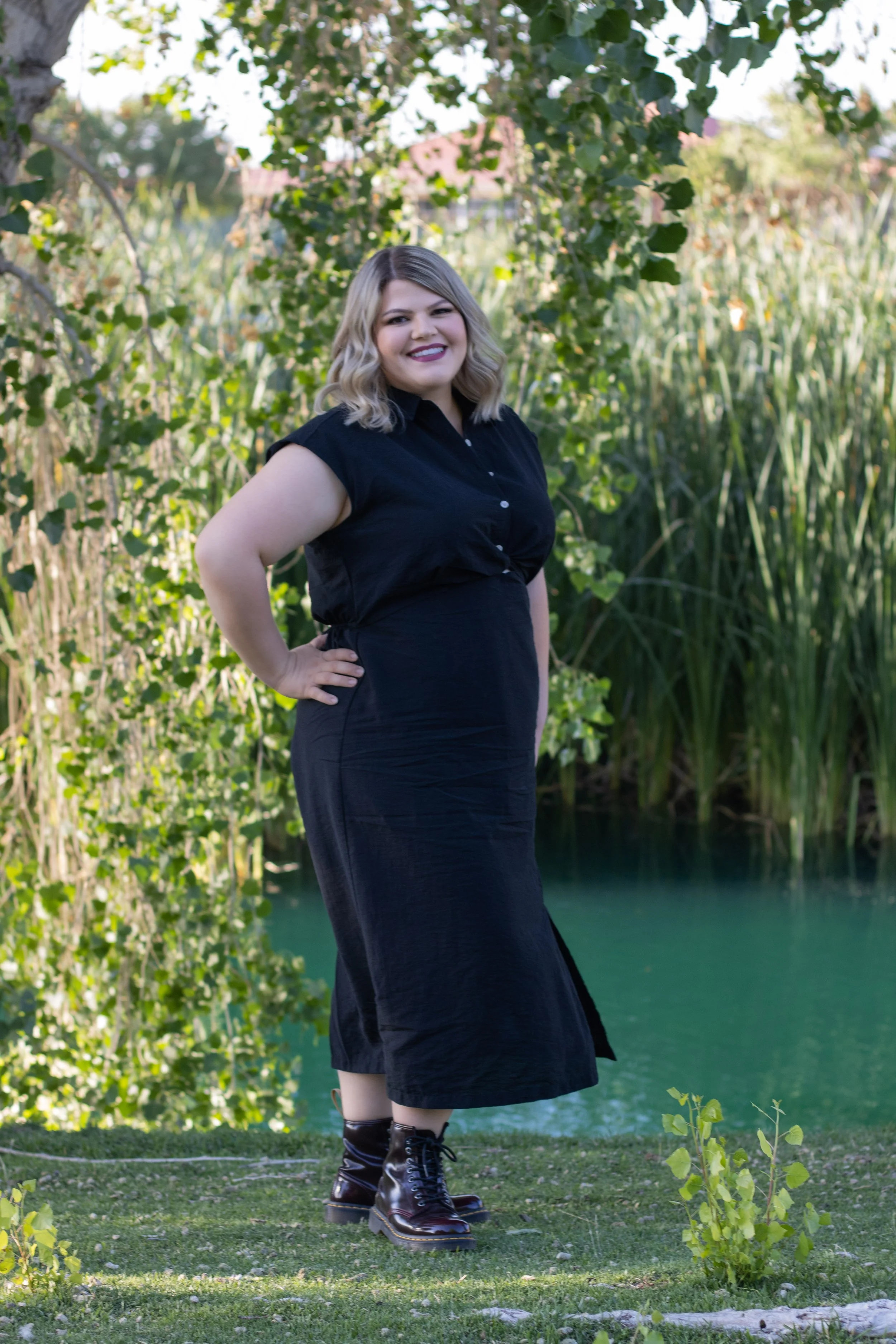Woman wearing black dress and doc martens smiling for graduation photos in front of lake by pinon hills photographer.