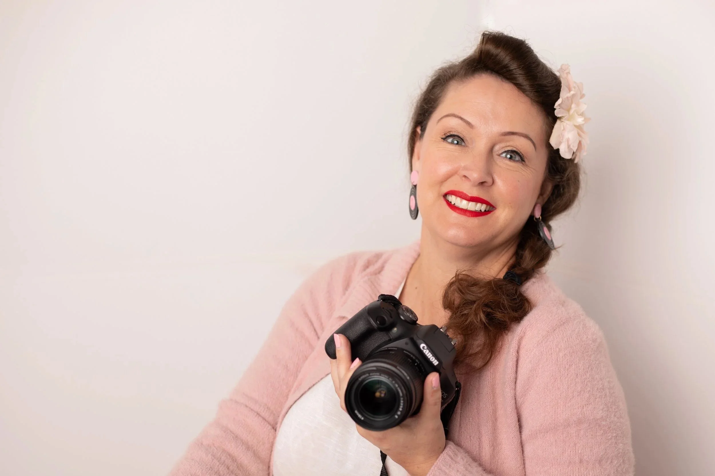 High Desert family photographer woman looking at camera and smiling wearing a pink sweater with flower in her hair. Holding a canon camera.