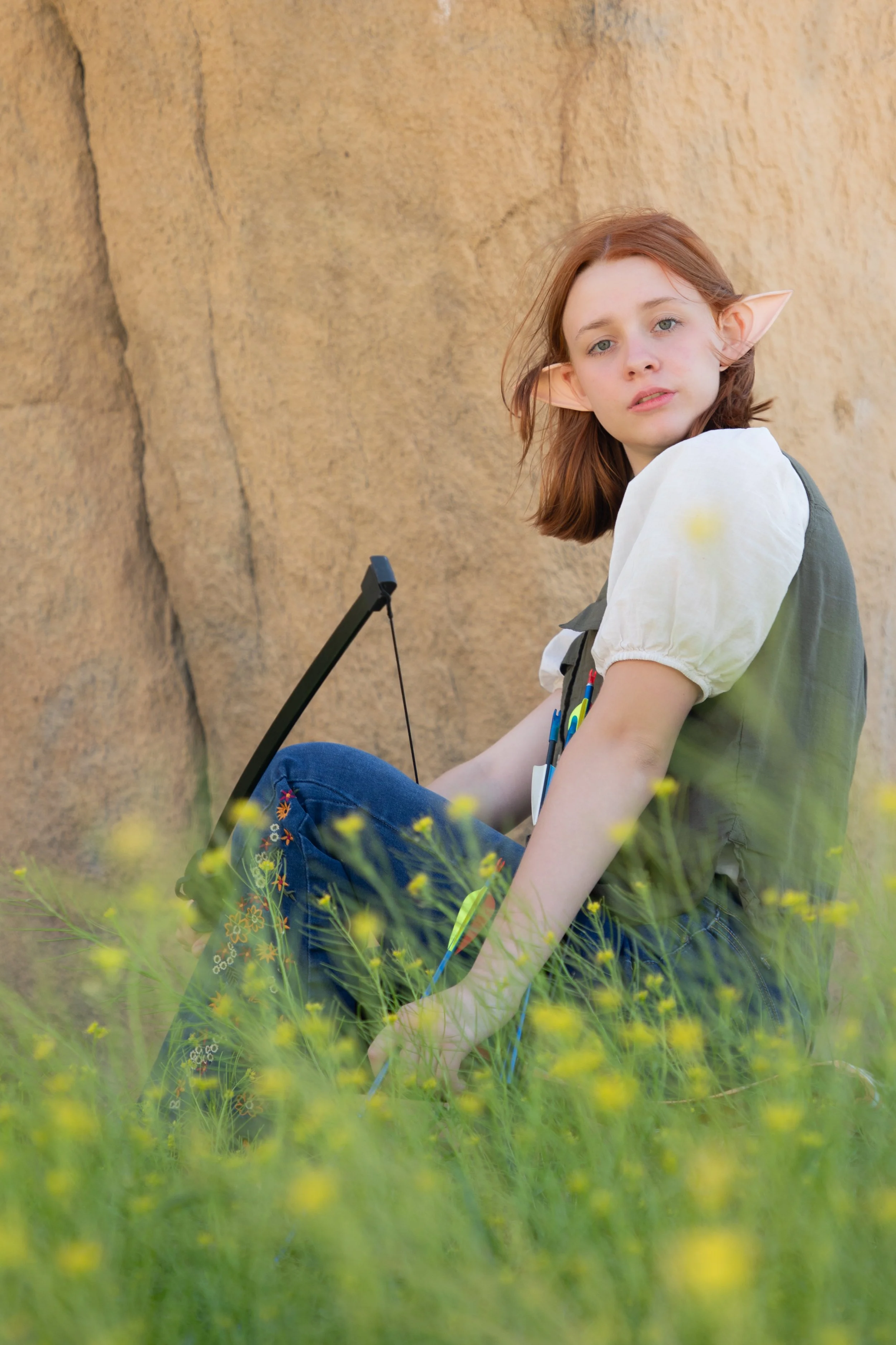 teen girl with elf ears holding bow and arrow sitting in the flower field