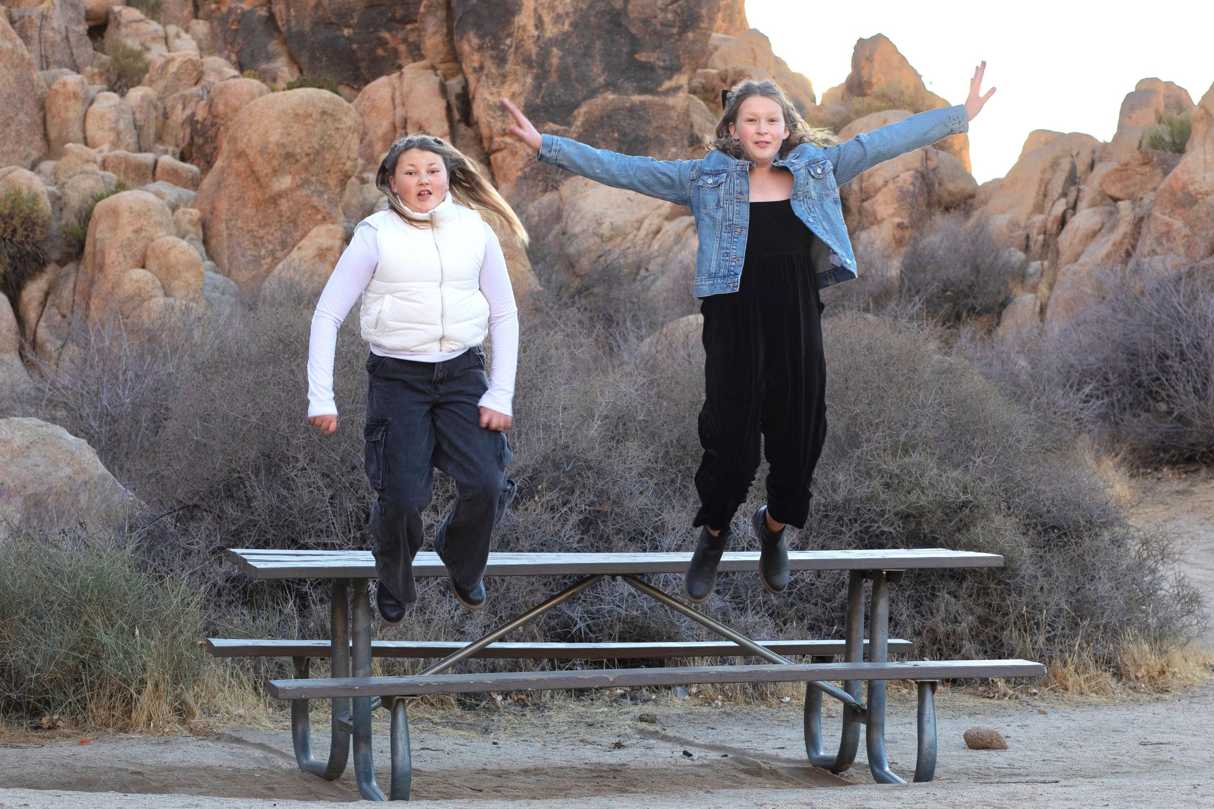 girls jumping off a picnic table near rocks. laughing and jumping with silly faces by playful family photographer.