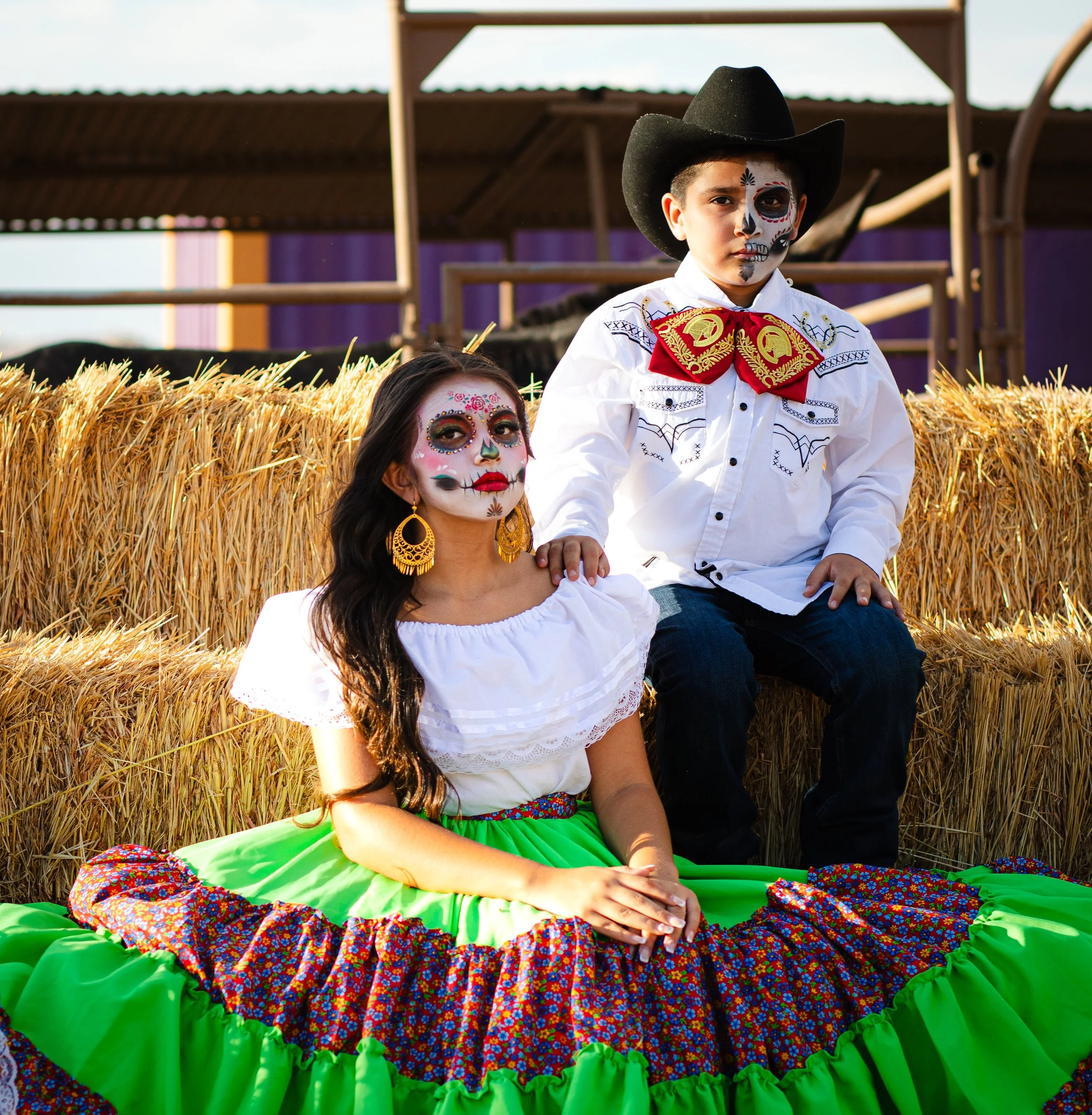 Brother and sister dressed for Dia de Los Muertos, sitting on hay bales.