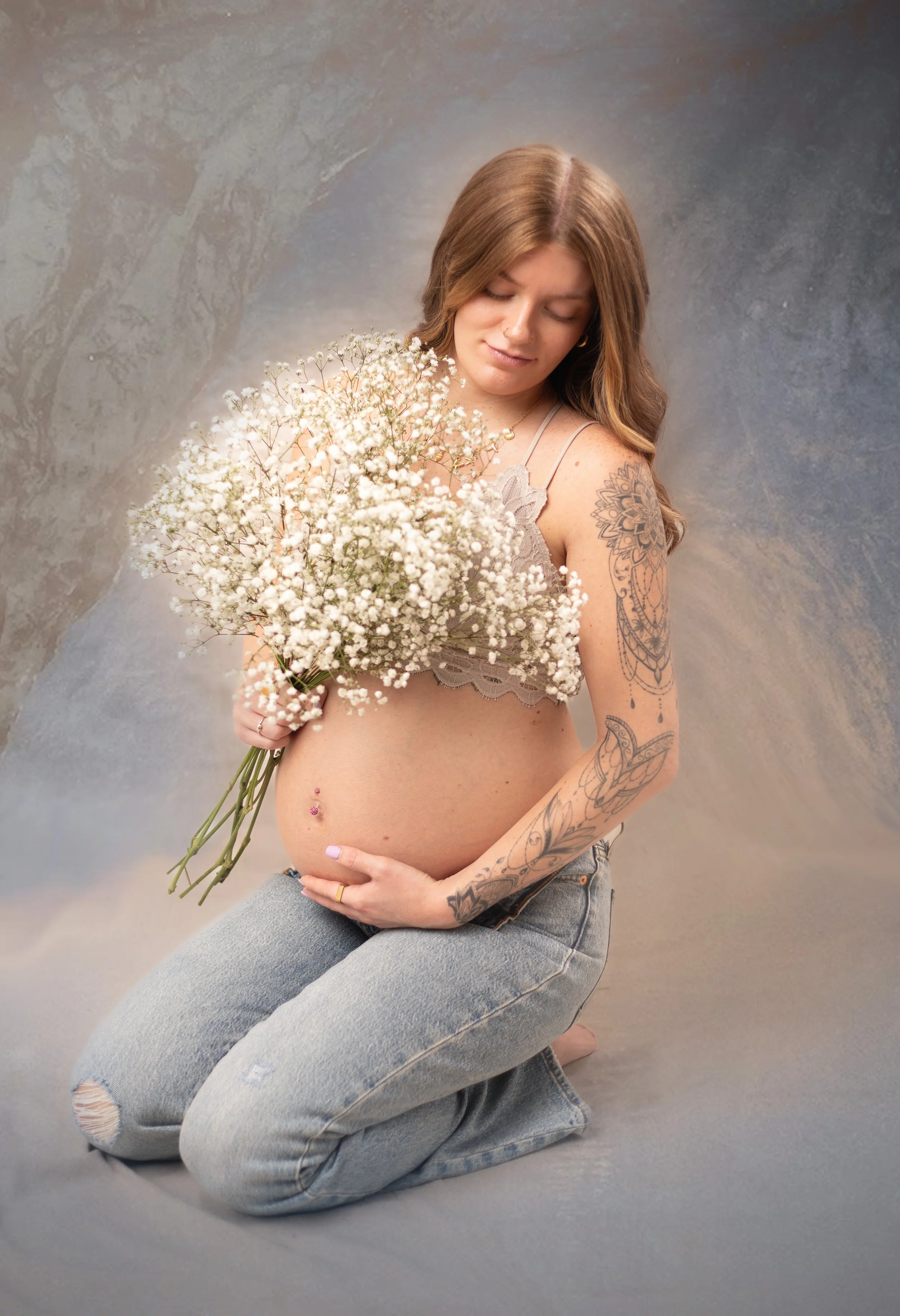 Simple maternity photo of mom in jeans holding flowers in studio, smiling looking at her belly. Family photographer in pinon hills, ca.
