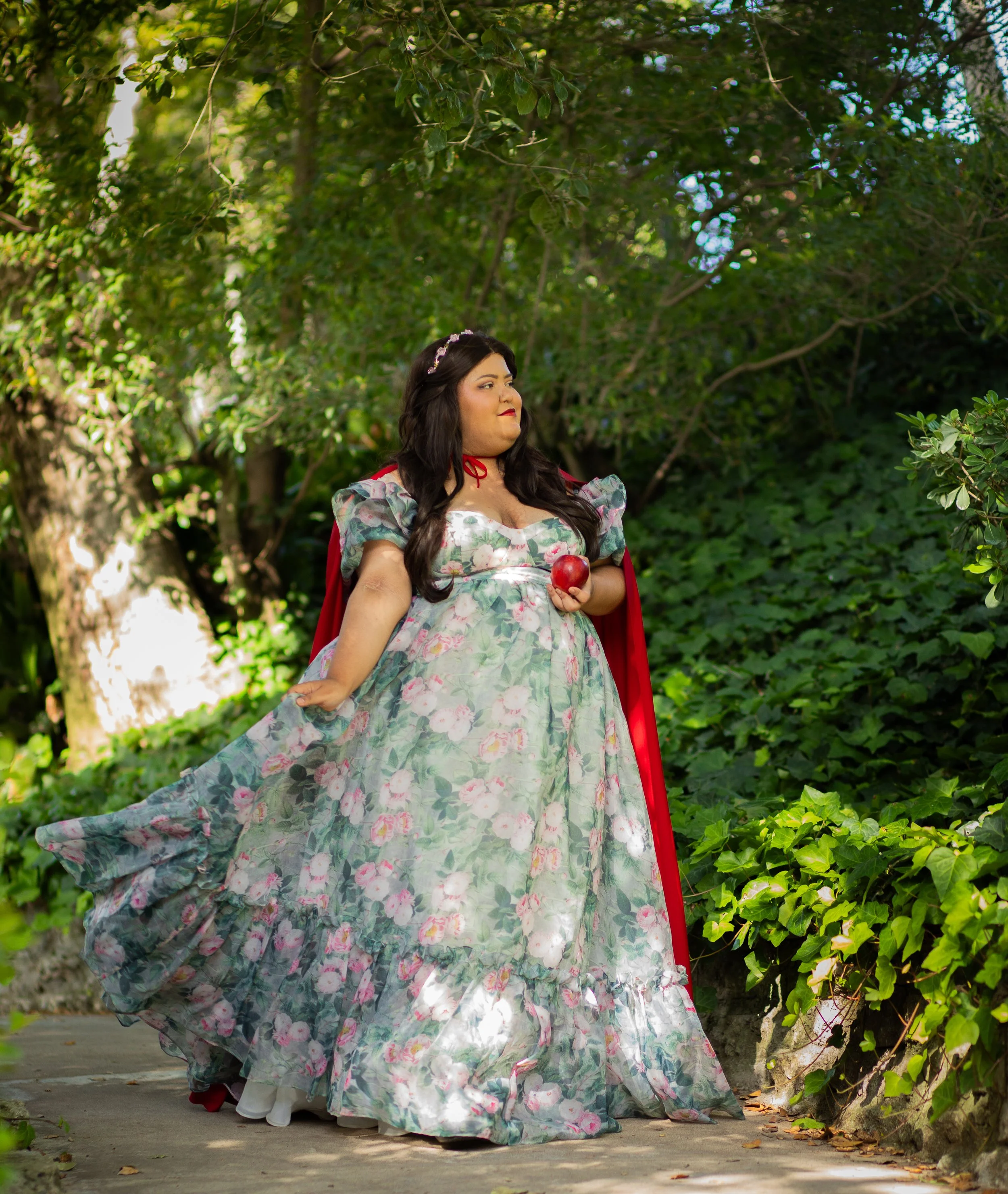 woman dressed as princess in green floral dress, red cloak, crown, holding apple and walking through the garden. Fantasy photographer in inland empire and high desert, southern ca.