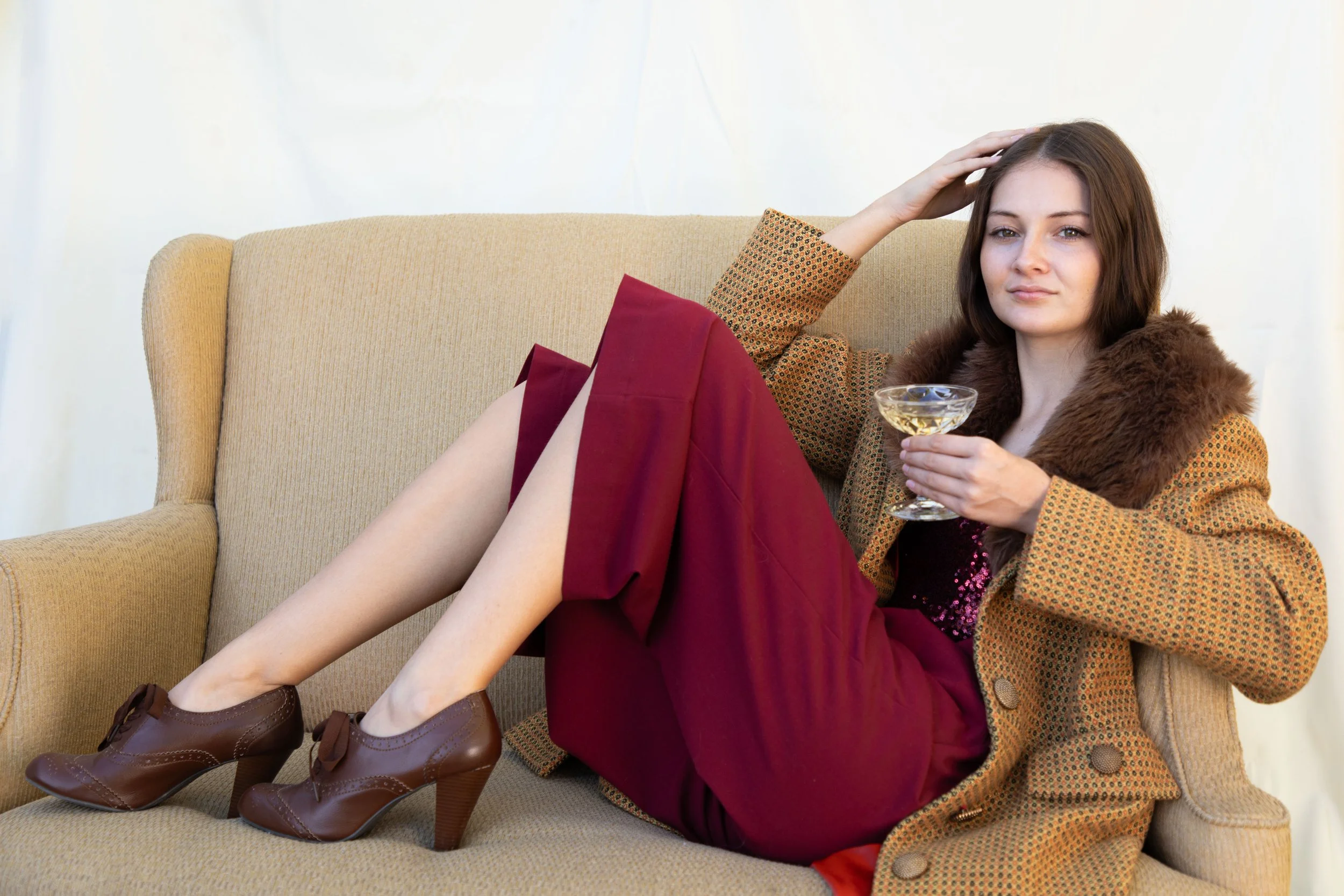 studio photo of woman sitting on couch wearing vintage clothing holding champagne glass, touching hair with other hand. Southern California fantasy photography studio sessions.