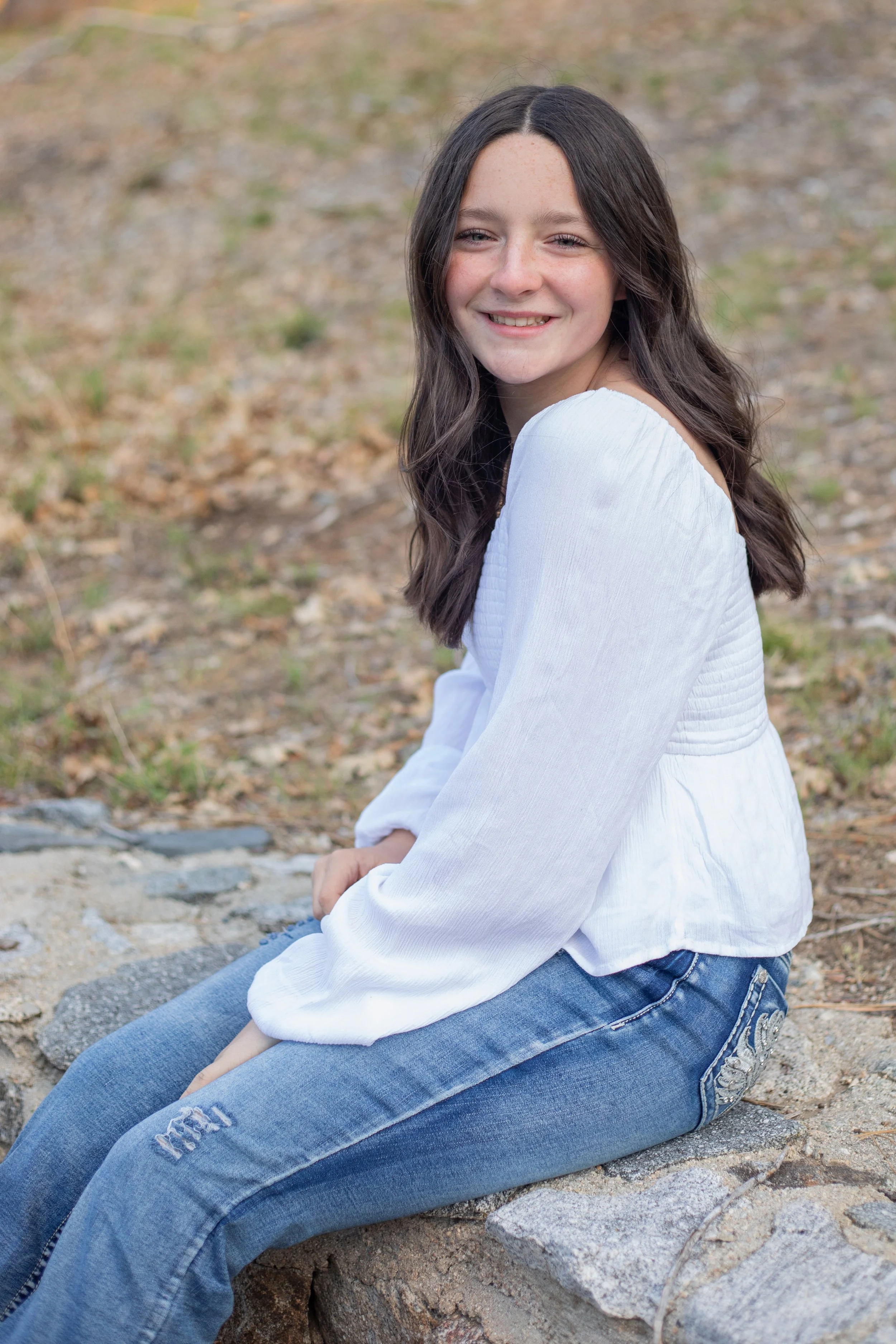 A teenage girl with long dark hair, wearing a white long-sleeve blouse and ripped blue jeans, sitting outdoors on a rock with a blurred natural background, smiling at the camera. Wrightwood family photographer.