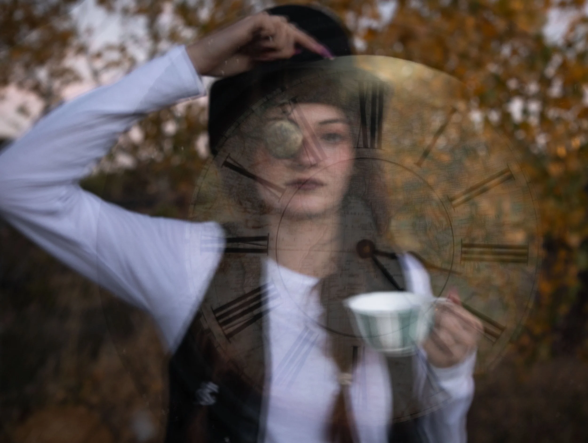 Woman dressed in steampunk holding pocket watch in front of eye, holding teacup in other hand. Outdoors, autumn trees, with clockface in front of person. Wrightwood fantasy photographer, Annette Marie creates magical worlds.