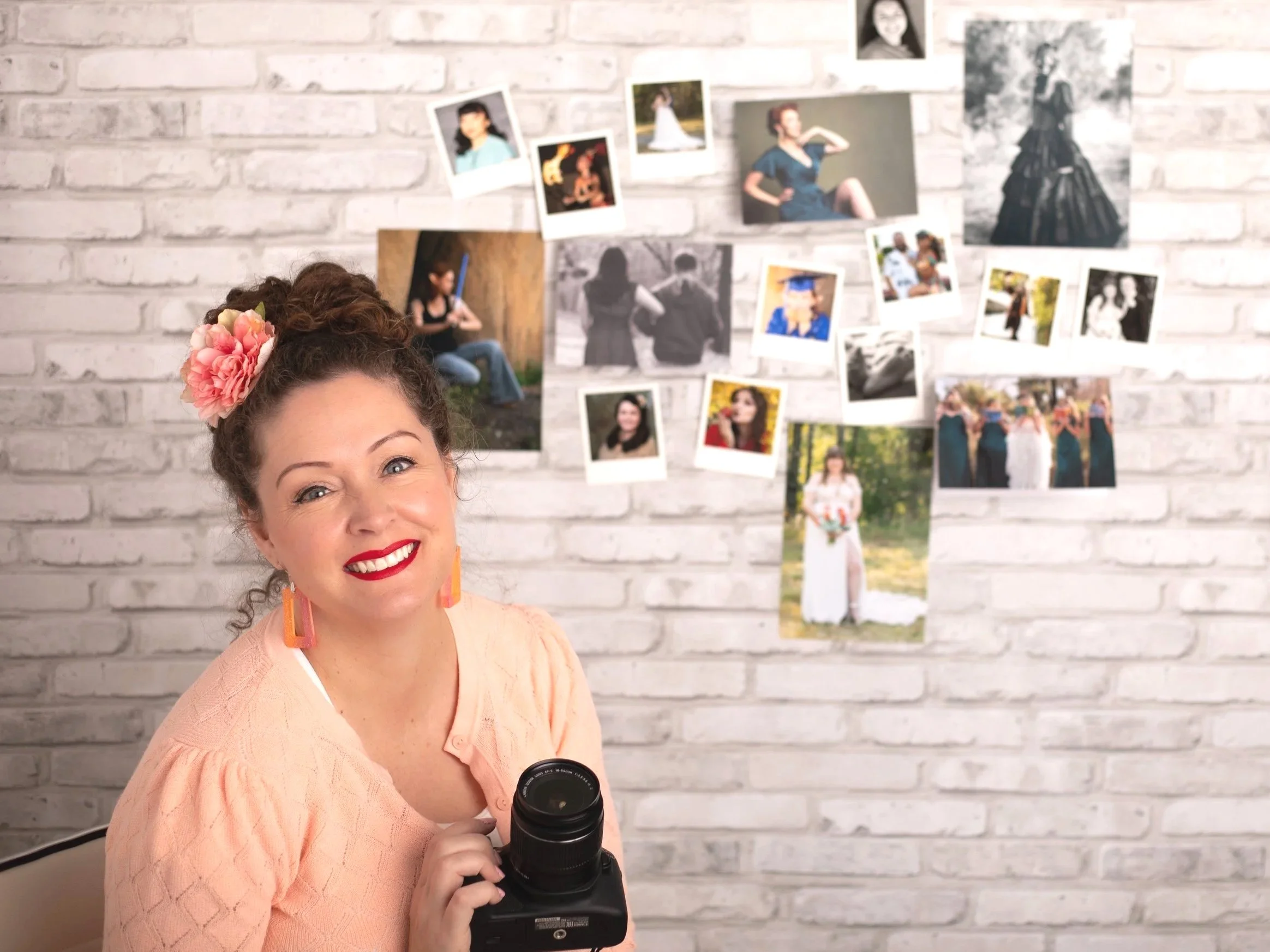 phelan family photographer woman smiling, wearing pink sweater, flower in hair, holding camera while sitting in front wall of printed photos.