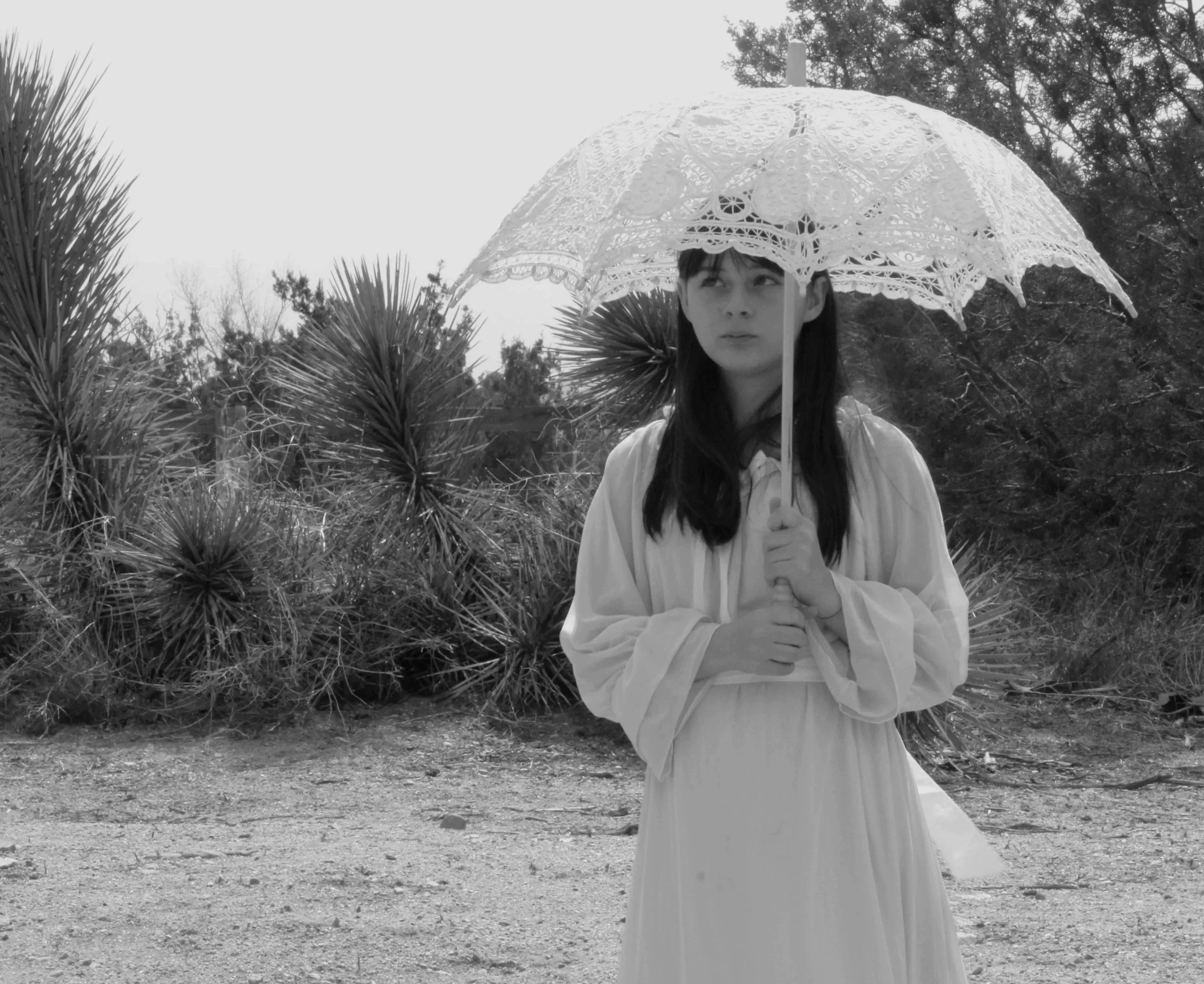 A young girl with long dark hair holding a white lace parasol outdoors. She is dressed in a light-colored dress, standing on a dirt path with Joshua trees in the background. The image is in black and white. Victorville Fantasy photography.
