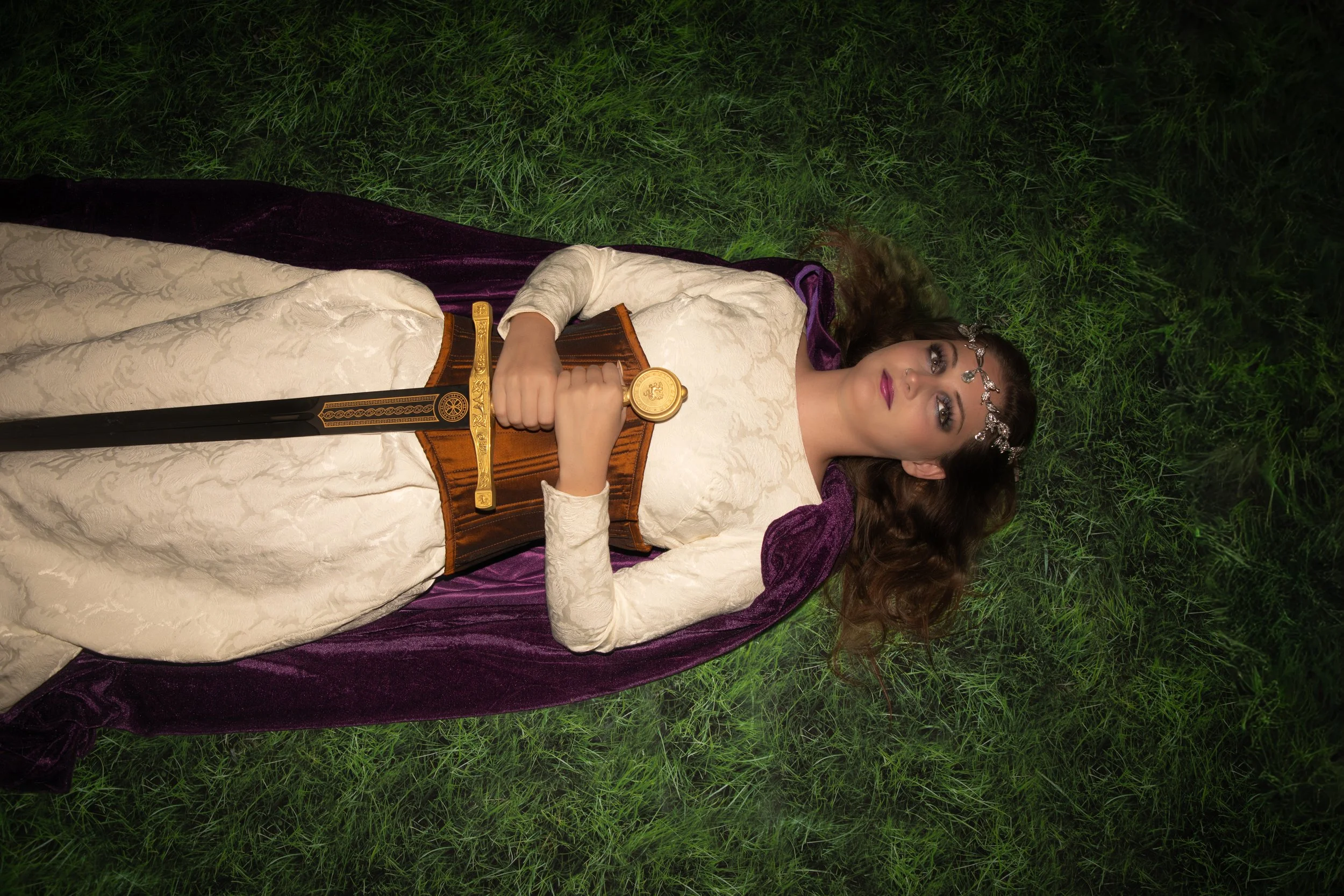 Woman in dress and corset, laying down in grass, holding sword. Studio sessions are a better choice than outdoor photography.