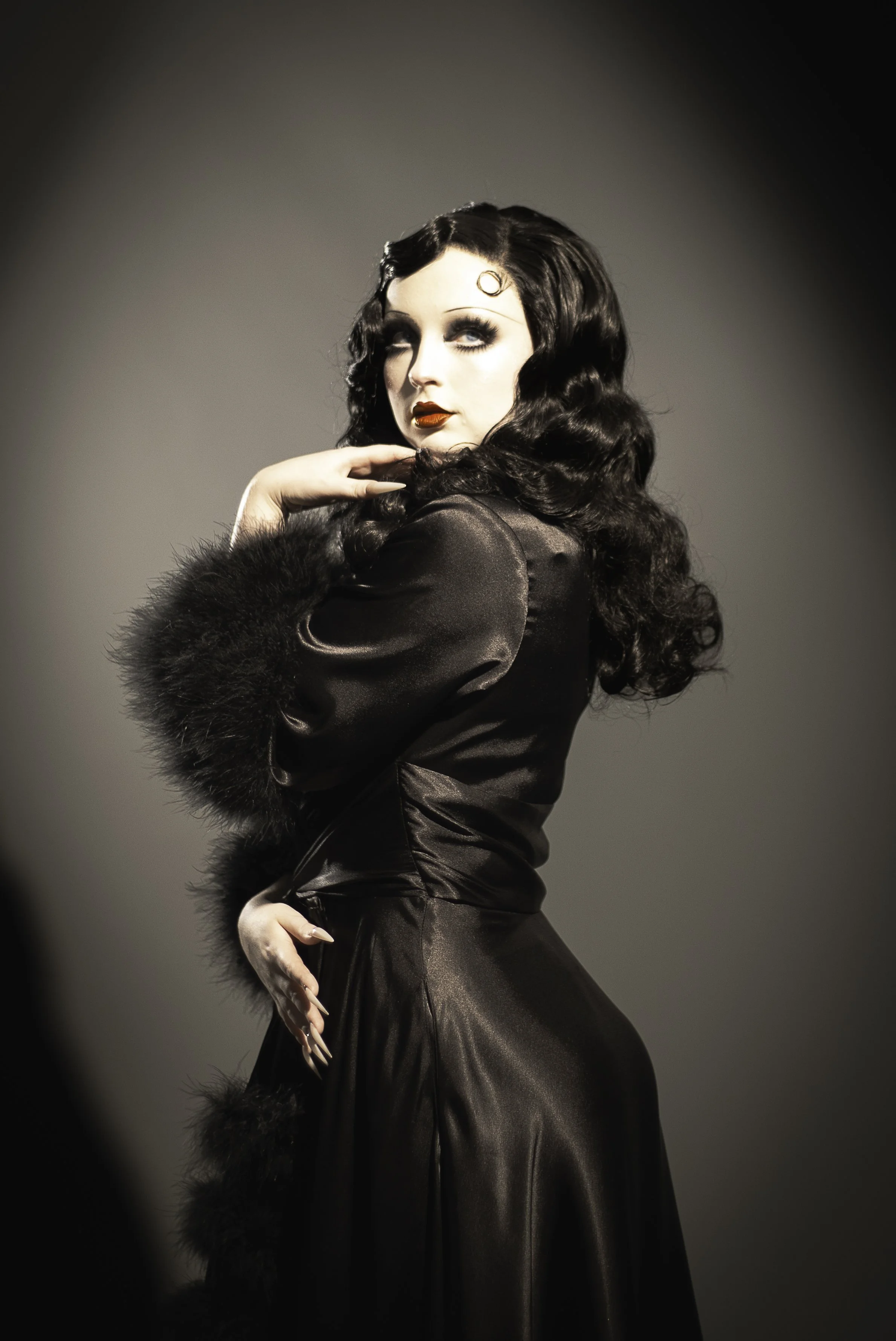 A woman with long, curly black hair and vintage makeup, wearing a black satin gown with fur trim on the sleeves, poses with her hand near her face against a neutral backdrop.