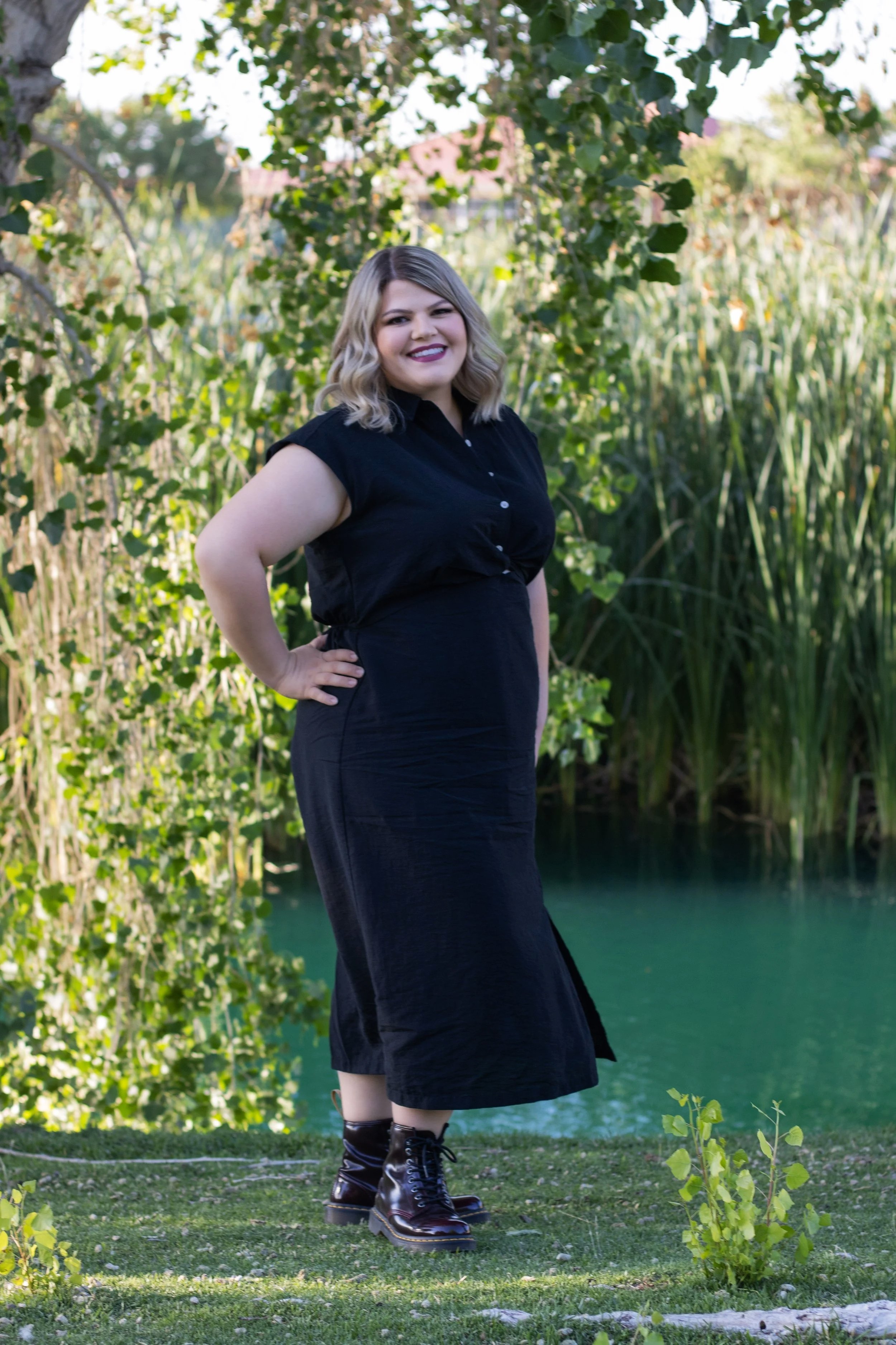 A woman with blonde hair and purple lipstick standing outdoors Victor Valley College near water, surrounded by trees and grass, wearing a black dress and boots, smiling at the camera, by fantasy photographer in victorville, CA.