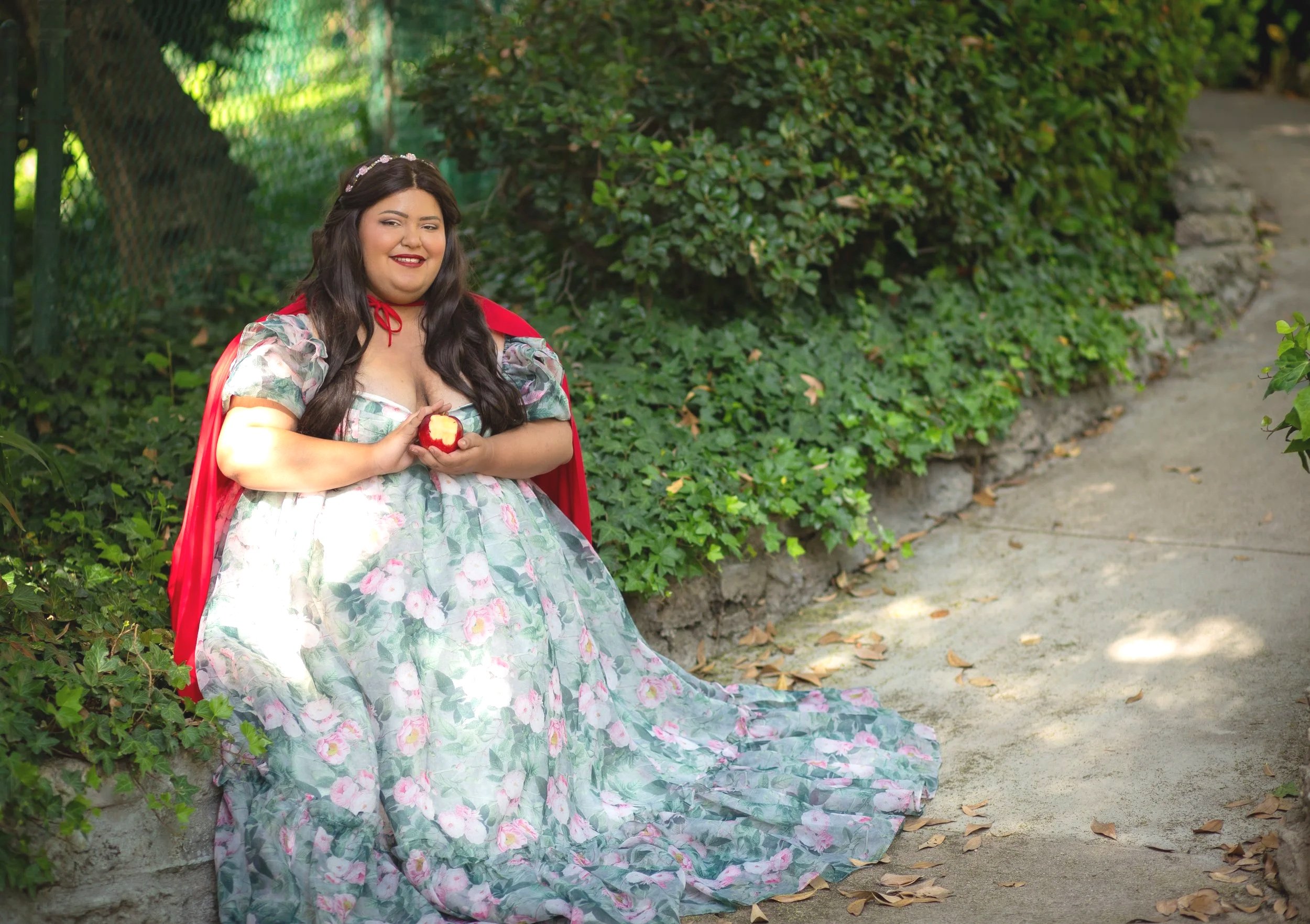 A woman dressed as Snow White sitting on a stone ledge outdoors, holding a red apple with a bite taken out, surrounded by green bushes and trees.