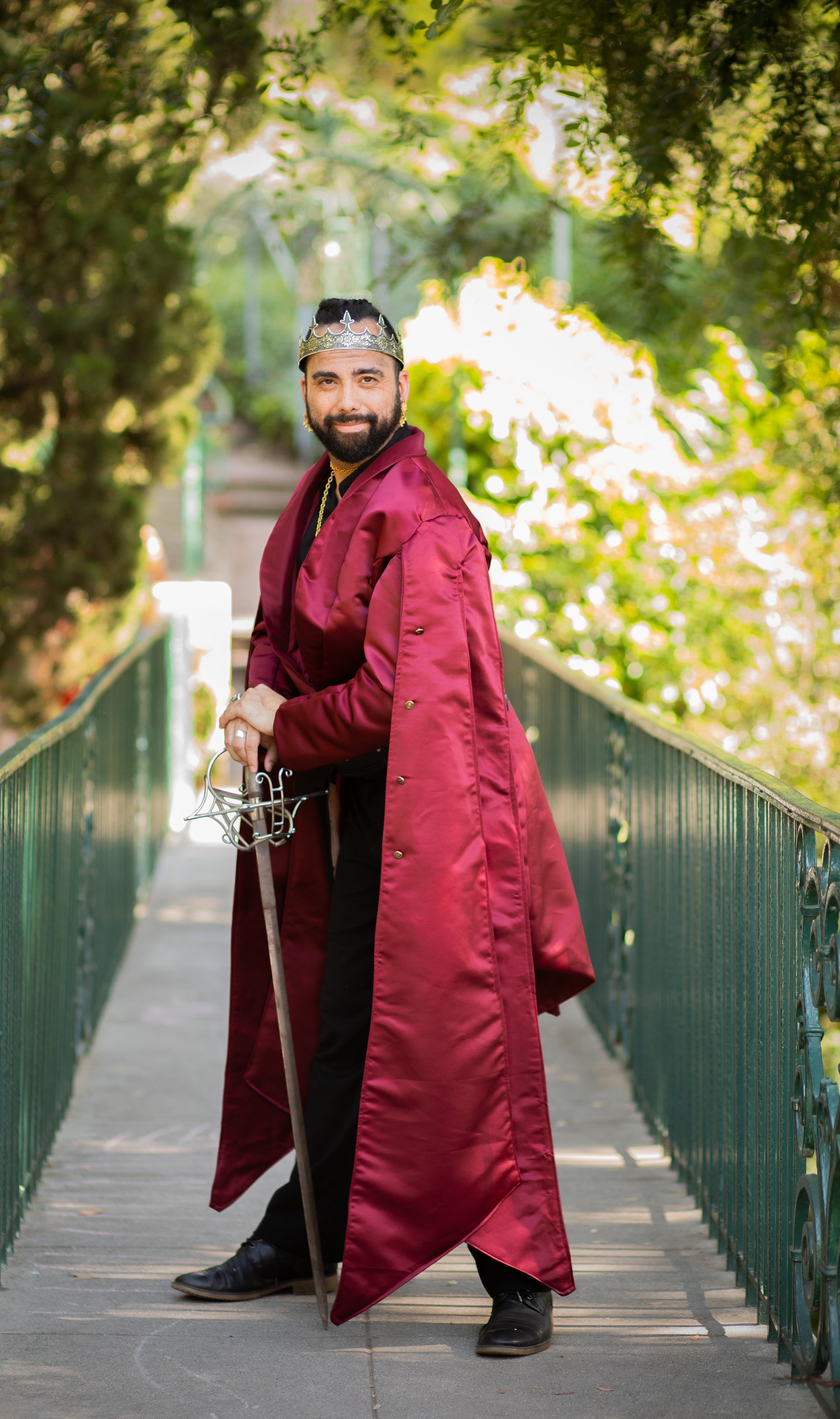 Man dressed as king cosplay holding sword, wearing a crown and red cloak. Standing on a bridge outside. Victorville photographer for fantasy and whimsy.