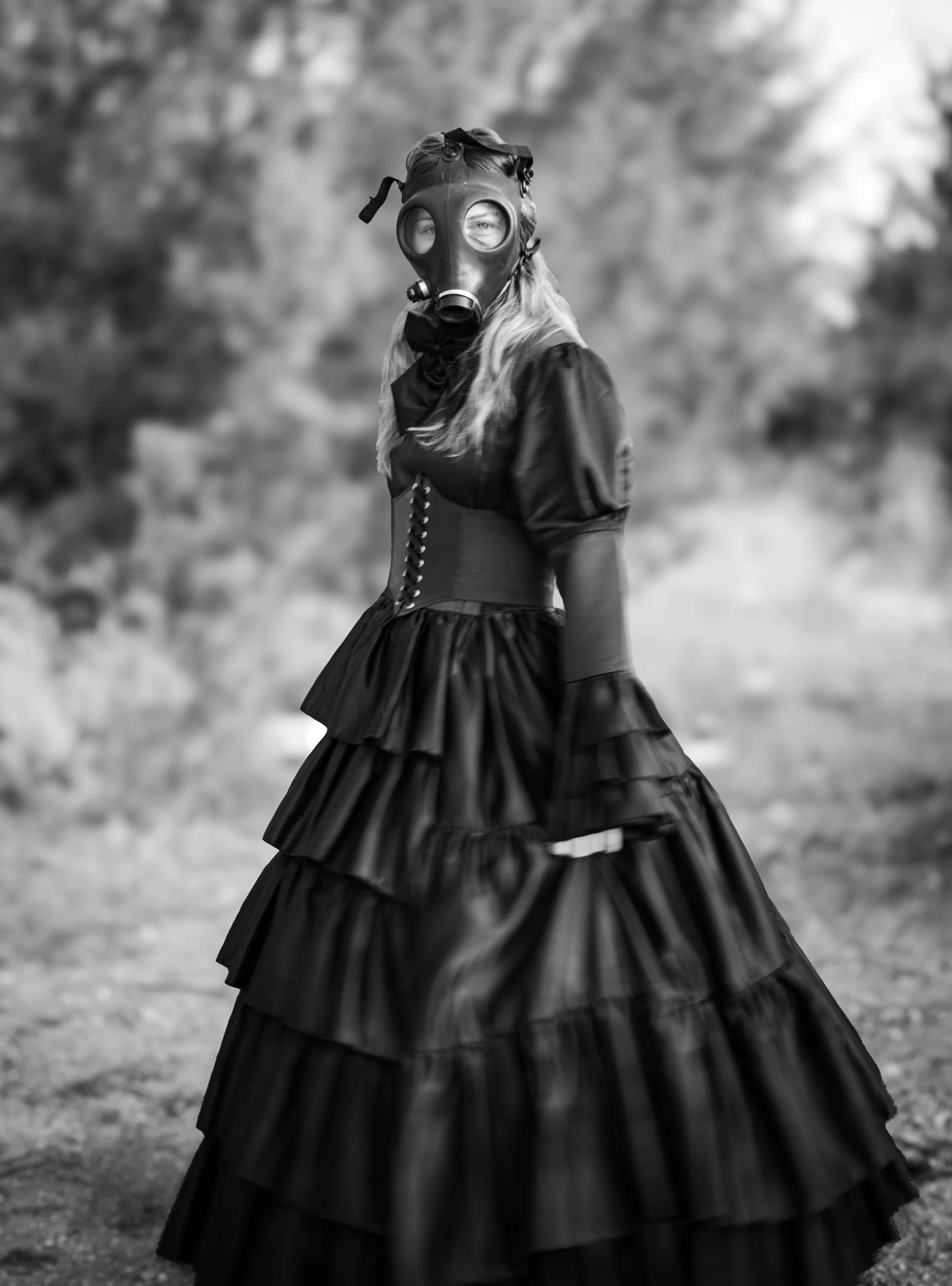 A woman in a long, layered, ruffled black dress and long sleeves wearing a gas mask, standing outdoors with a blurred natural background.