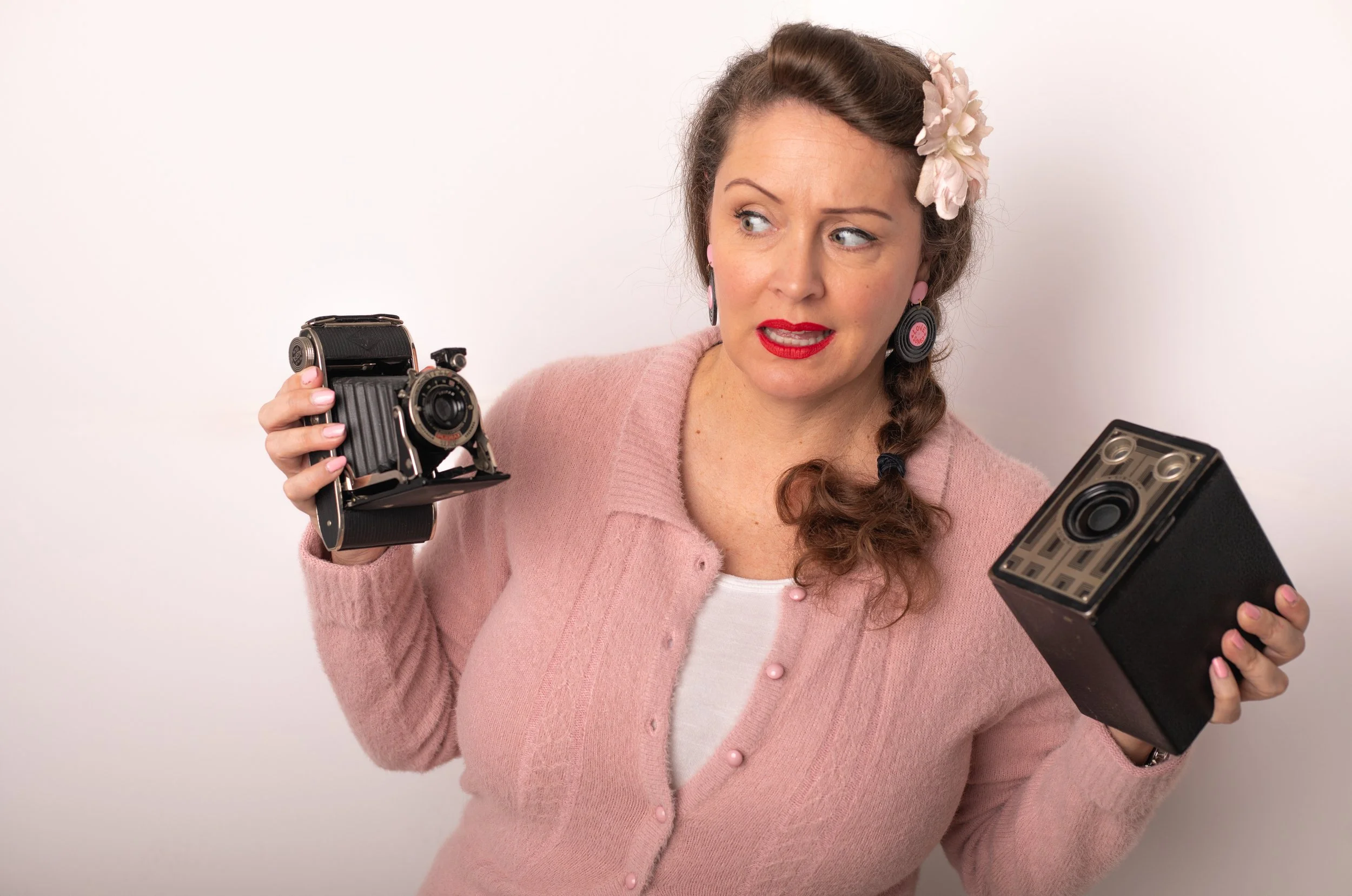 pinon hills photographer holding vintage cameras looking confused at which one to use. woman wearing pink sweater and side braid.