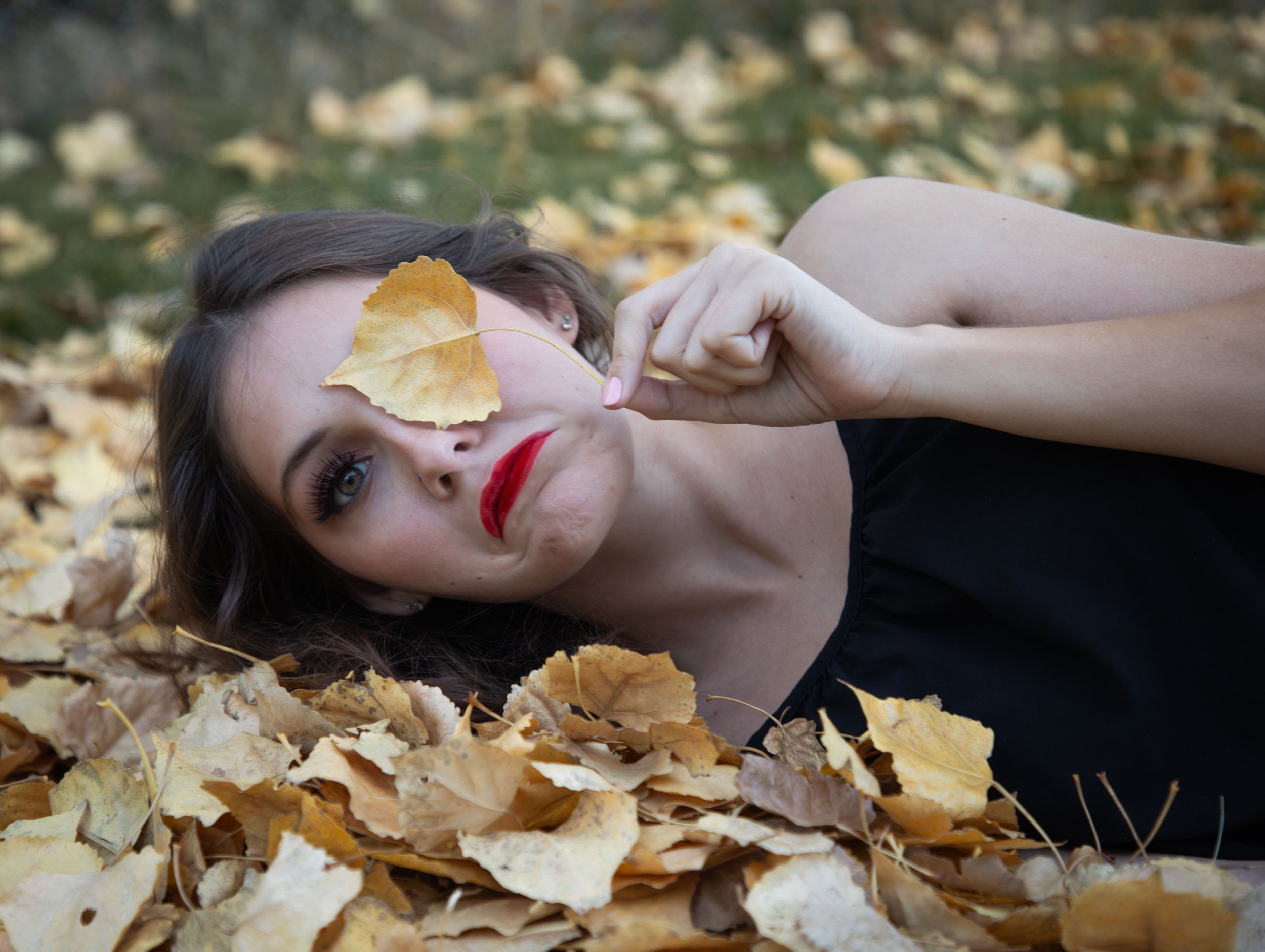 Woman laying in a pile of leaves with one leaf covering her eye, frowning.high desert fantasy  and family photography.