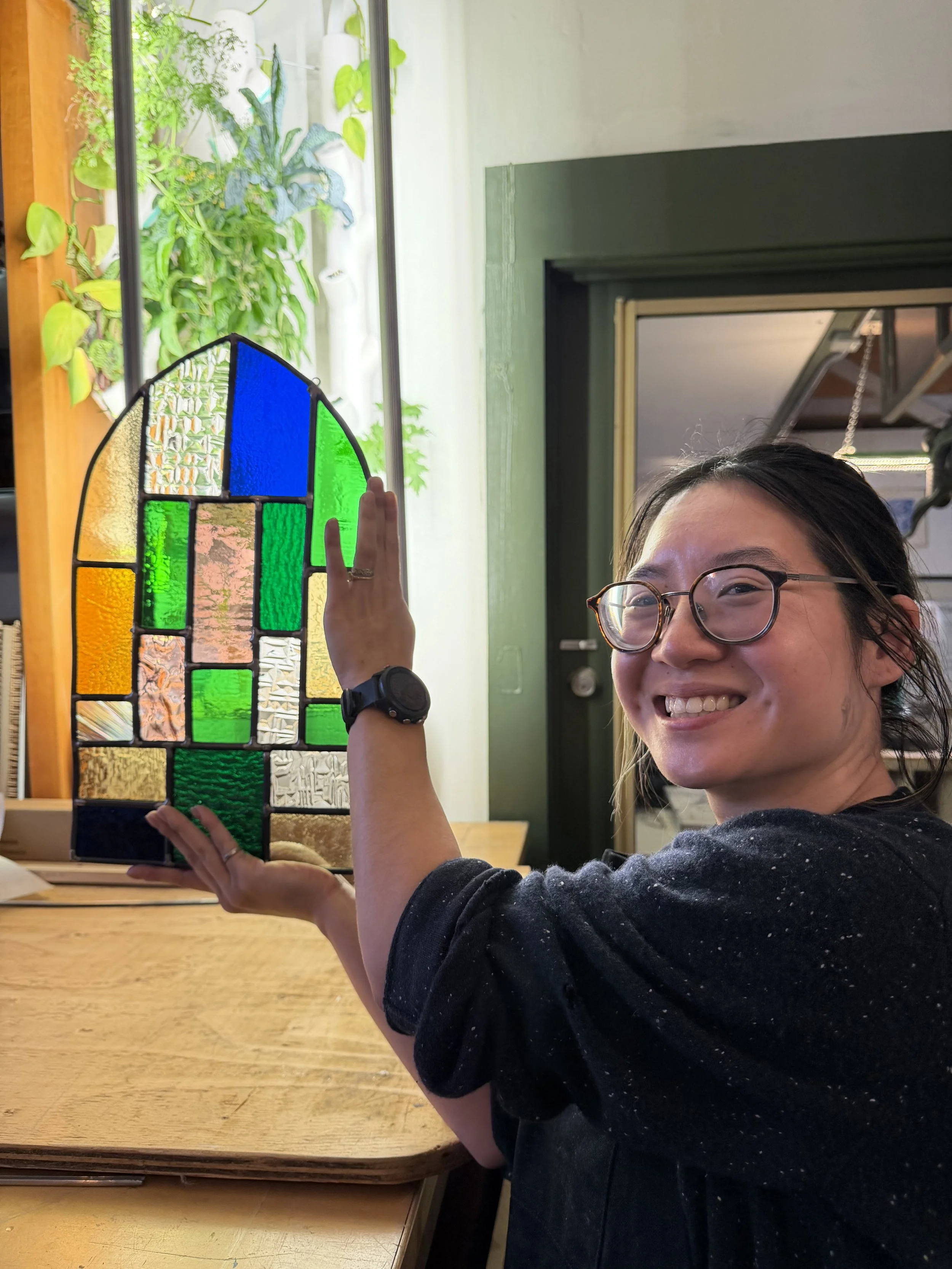 A woman with glasses smiling while holding a colored stained glass piece indoors with green plants in the background.