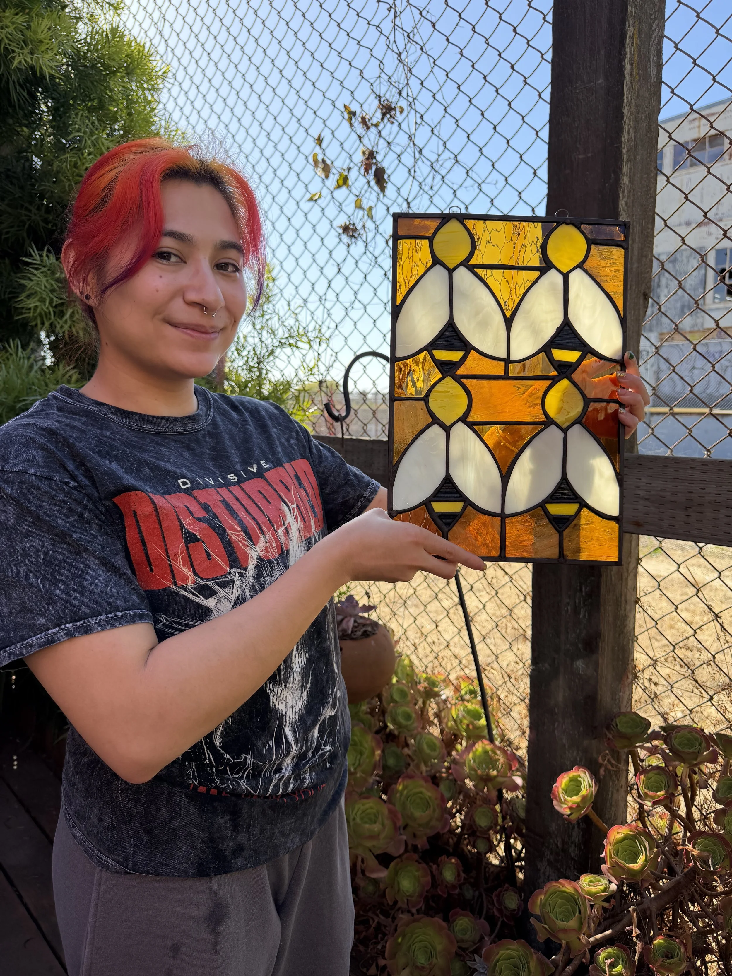 A young woman with colorful red and orange hair, smiling, holding a stained glass artwork with yellow, white, and black patterns outdoors near a chain-link fence and green plants.