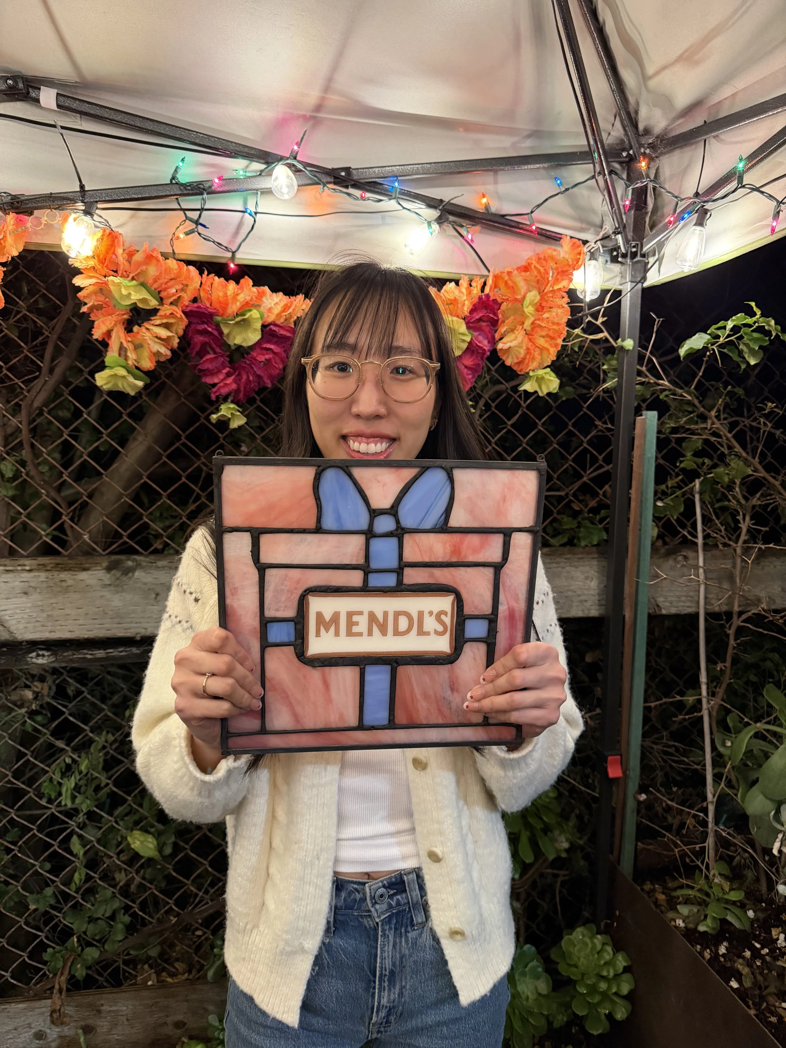 A smiling woman wearing glasses, a cream-colored cardigan, and jeans, holding a stained glass piece with a bow design and the word 'MENDL'S' in front of her. She is standing outdoors, with a fence, plants, and decorative orange and pink flowers in th