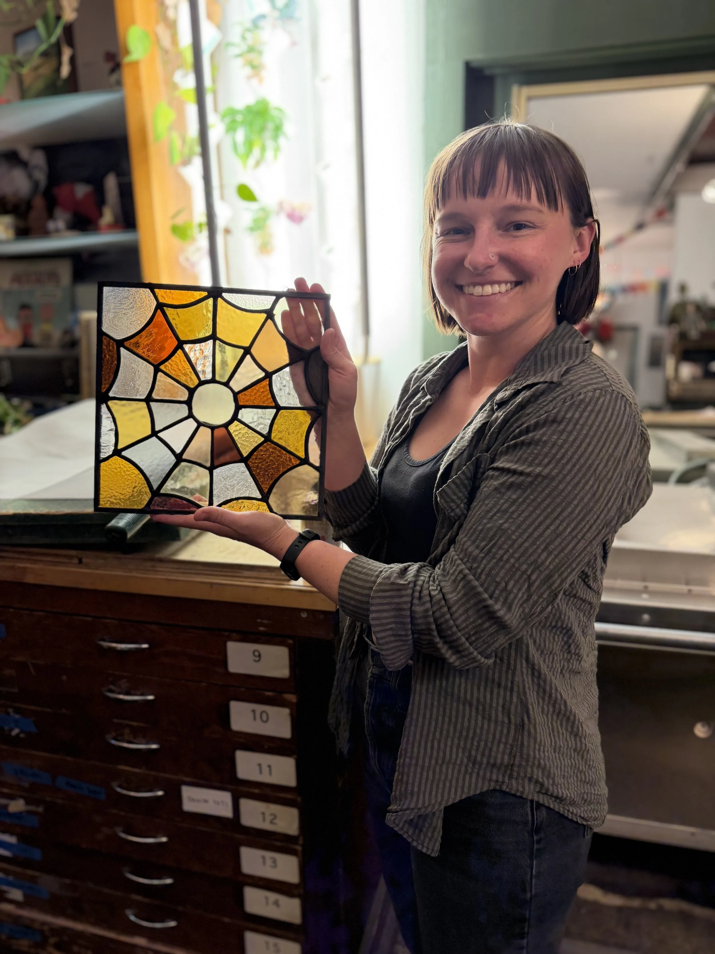 A woman with short brown hair holding a colorful stained glass window panel and smiling. She is wearing a striped shirt and stands in a room with shelves, drawers, and bright natural light coming from behind.