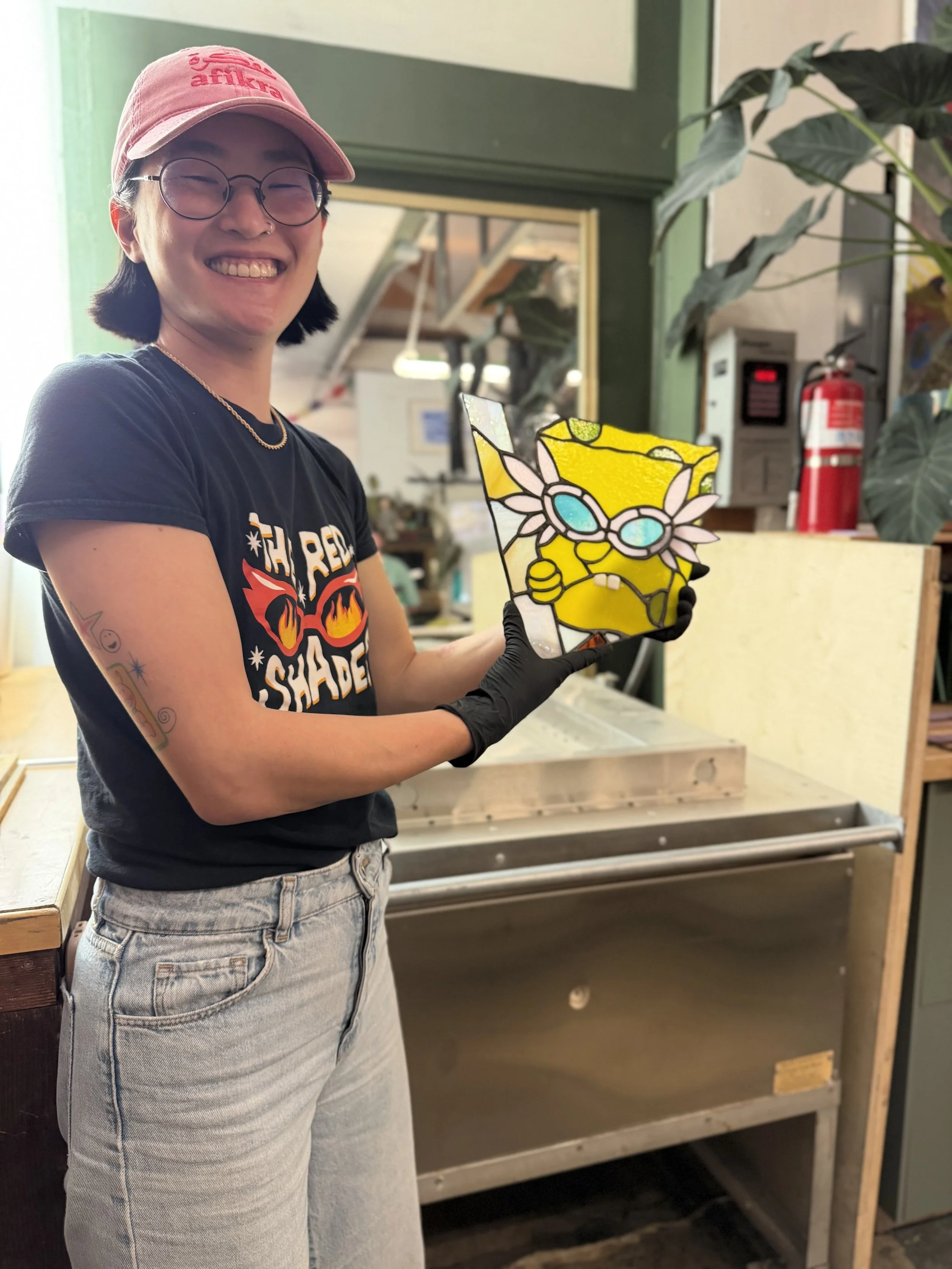 A person smiling while holding a colorful stained glass art of SpongeBob SquarePants, wearing a pink baseball cap, glasses, and a black T-shirt.