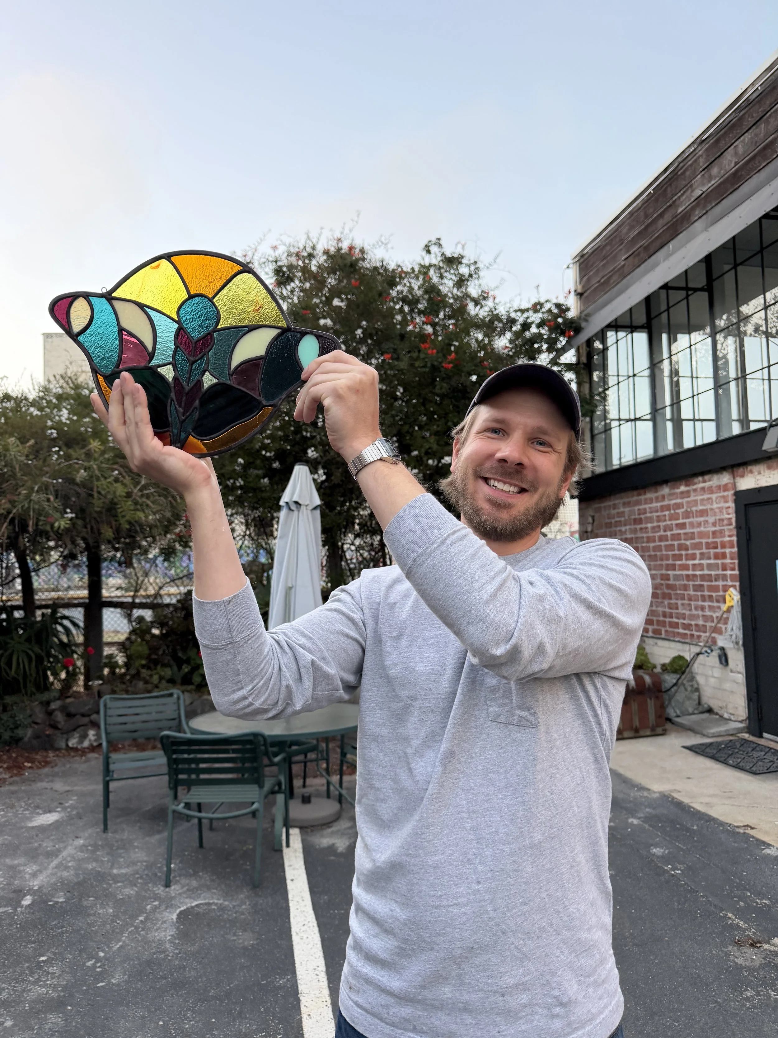 A man with a beard and wearing a gray shirt and cap is smiling and holding up a colorful stained glass art piece outdoors, with a building and trees in the background.