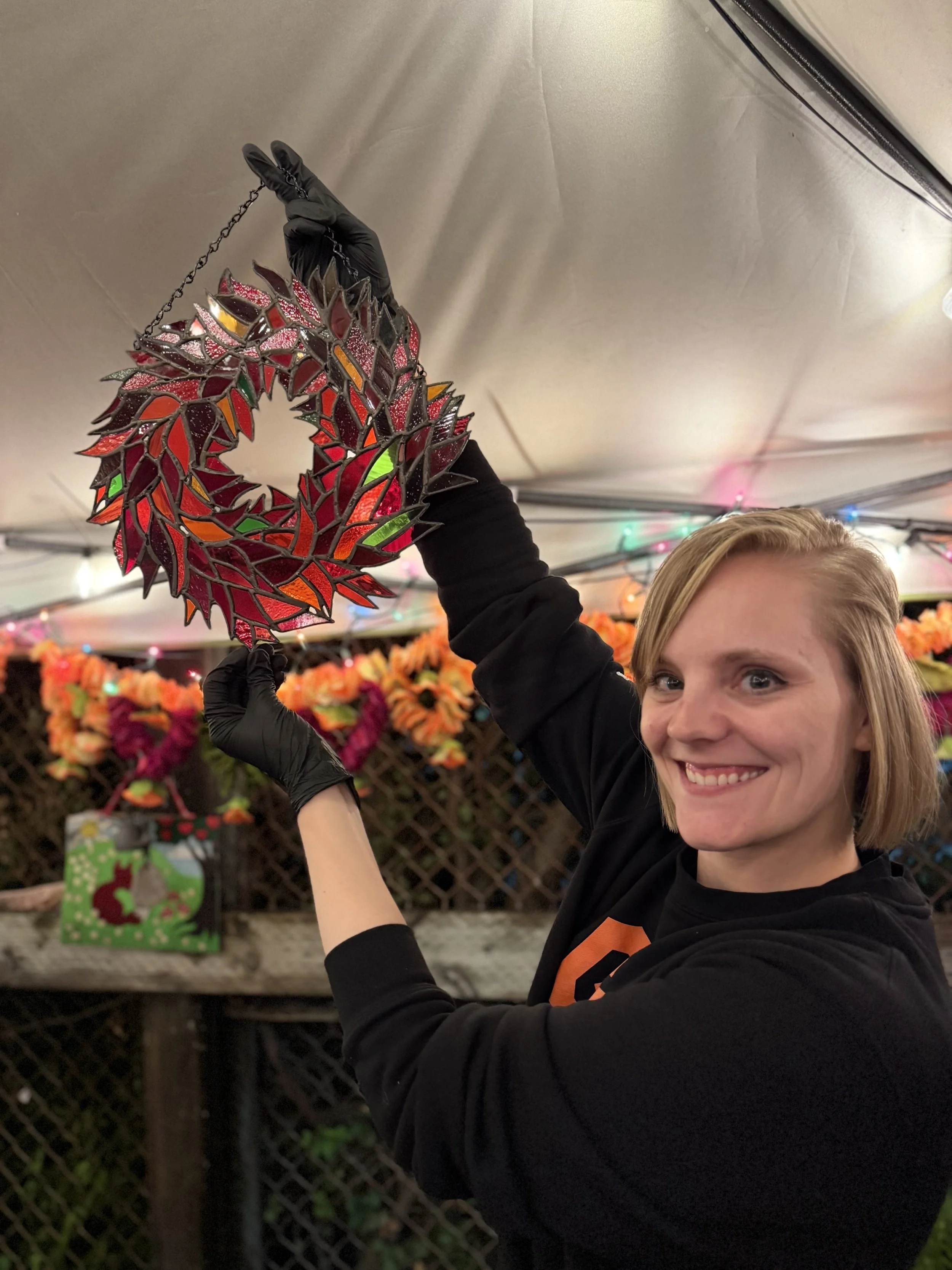 A smiling woman with blonde hair in a black shirt holding up a Christmas wreath made of stained glass. The background shows colorful holiday decorations and a gift bag with a reindeer design.