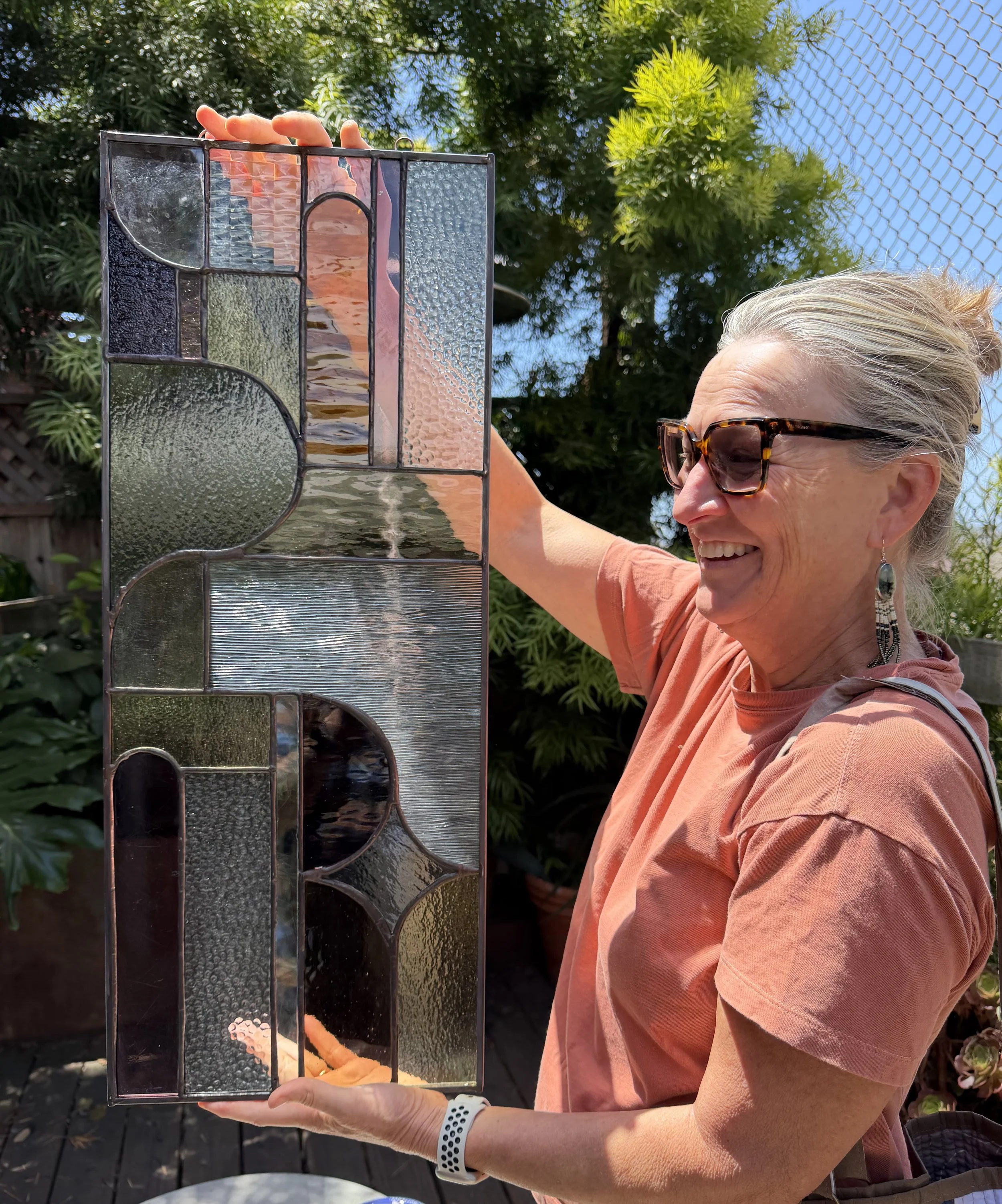 A smiling woman with sunglasses and earrings holding a stained glass panel outdoors. She is wearing an orange shirt and a white watch. A chain-link fence and green trees are in the background.