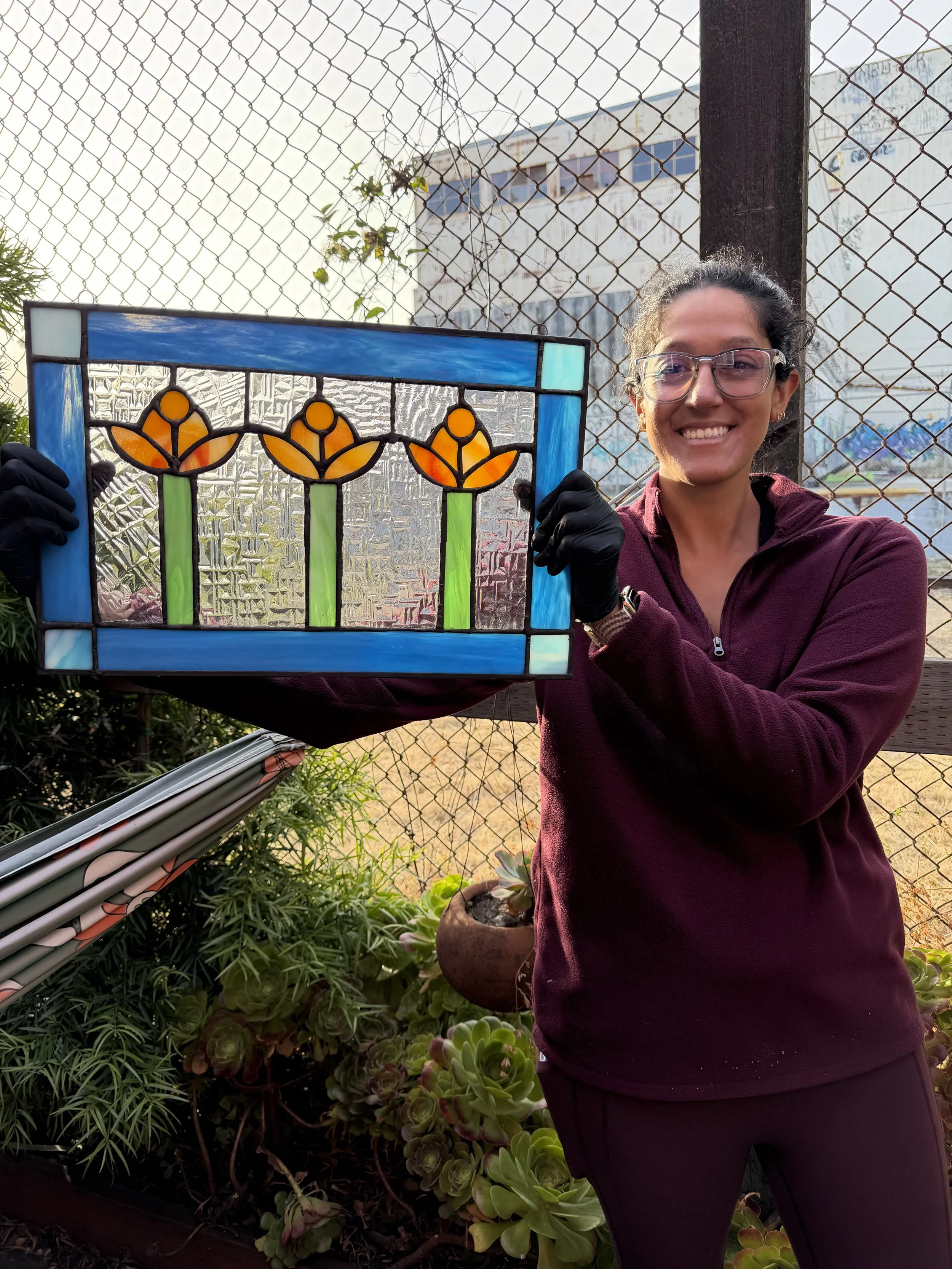A smiling woman wearing glasses and a purple jacket holds up a colorful stained glass artwork featuring three orange and yellow flowers with green stems, standing outdoors near a garden with various plants and a chain-link fence in the background.