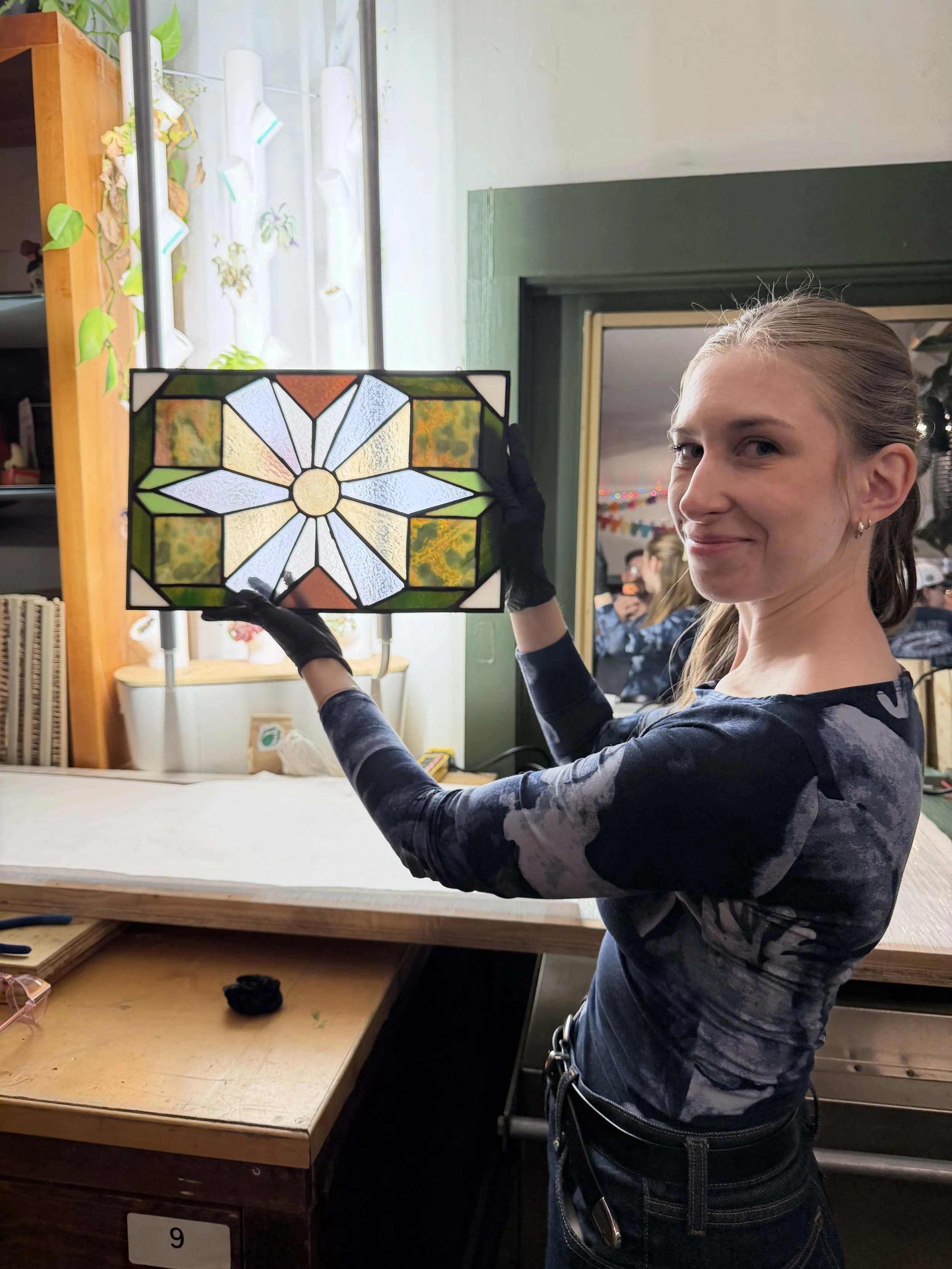 A woman with long hair tied back, smiling, holding a stained glass window panel in front of her. She is wearing a black and gray patterned long-sleeve shirt and black gloves. The background shows a room with a window, a mirror, and other people.