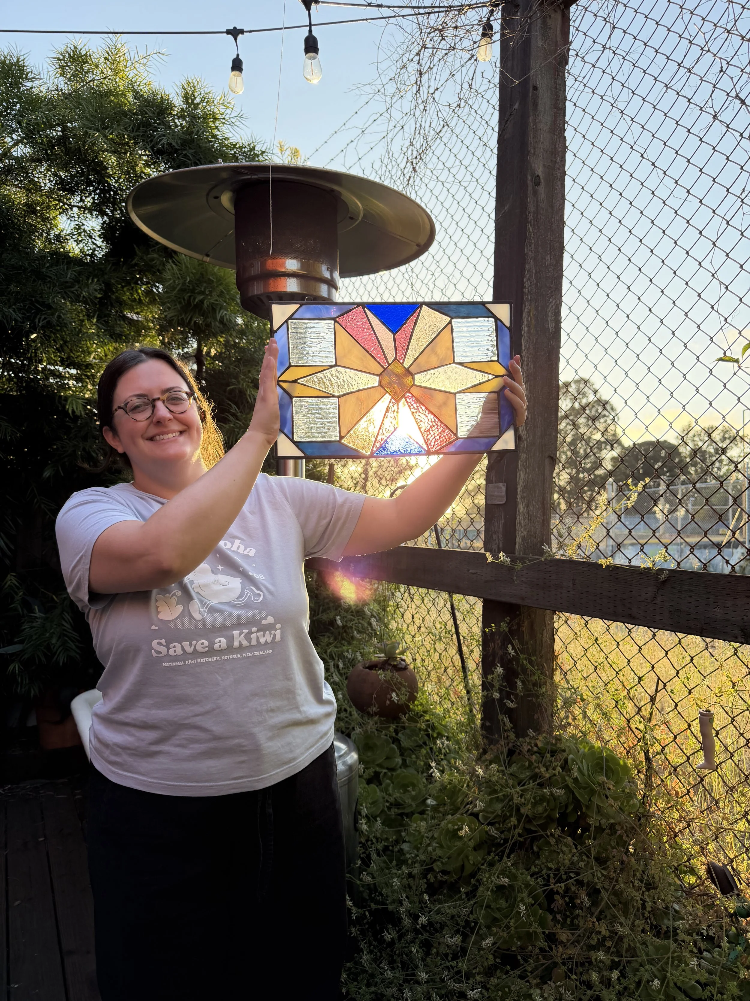 A woman smiling while holding up a stained glass panel with a geometric design, outdoors in front of a wooden fence and green plants, during sunset.