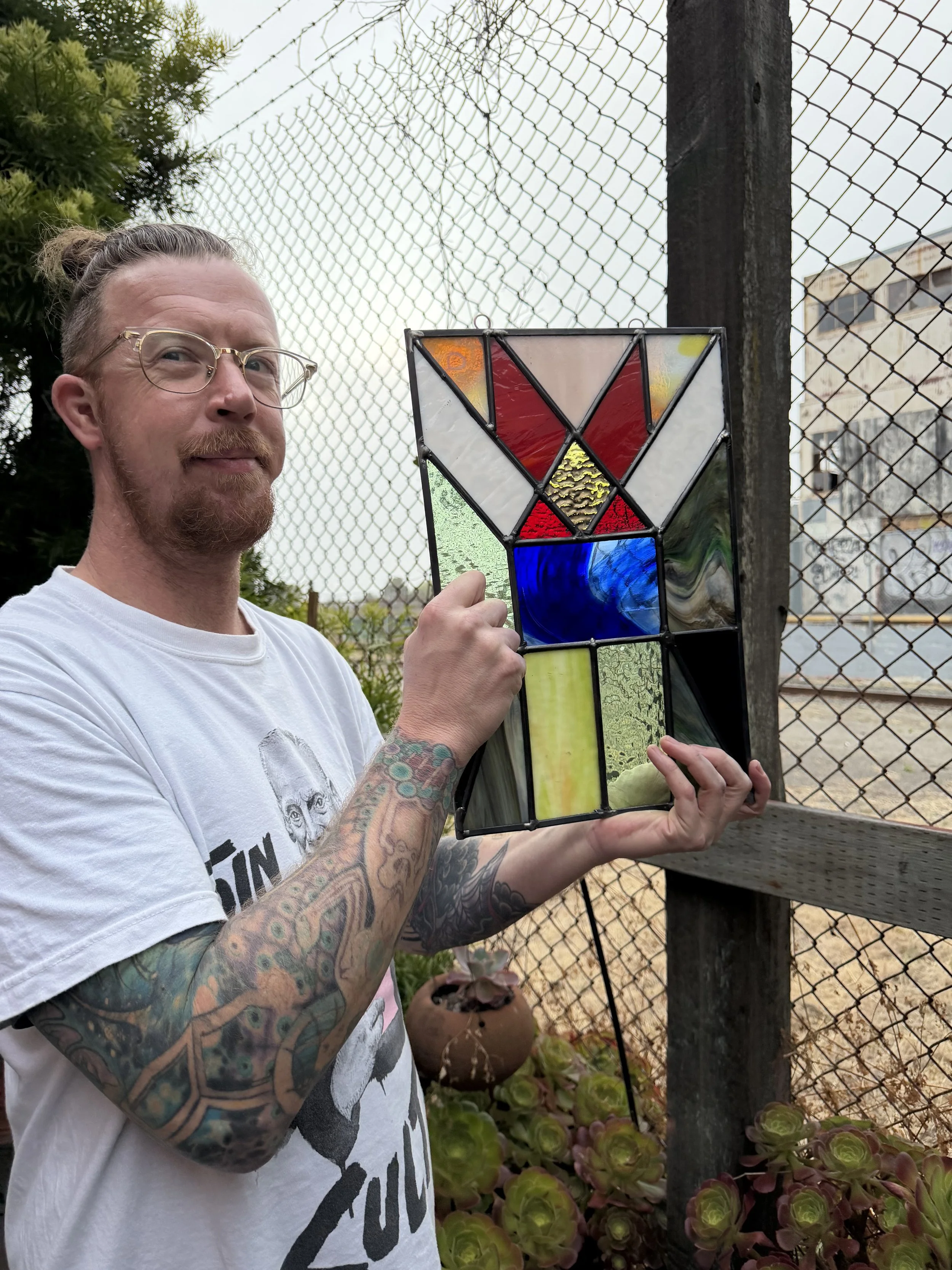 A man with glasses and tattoos holding a stained glass artwork in an outdoor setting next to a chain-link fence with plants and a building in the background.
