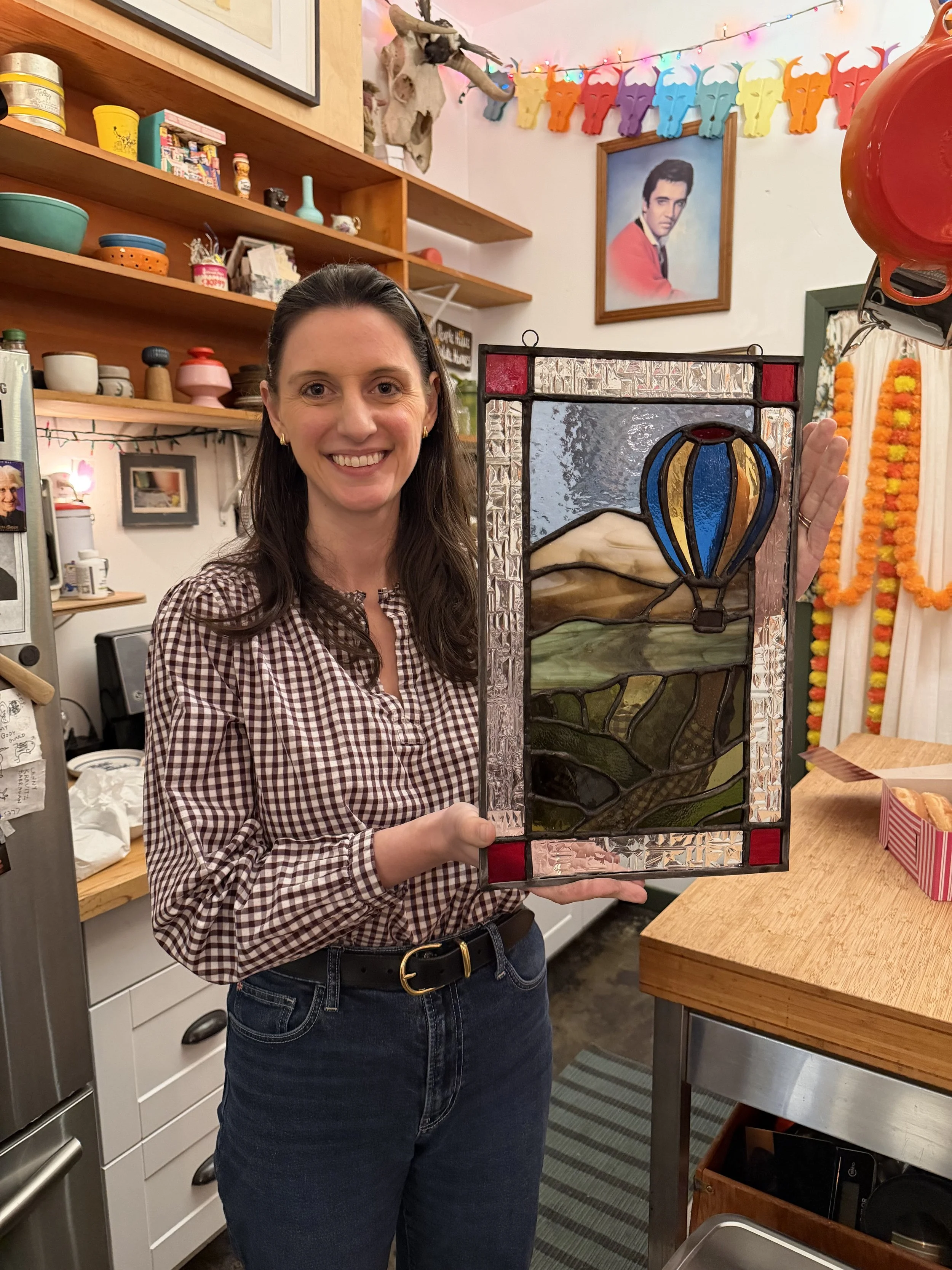 A woman smiling and holding a stained glass artwork with a hot air balloon and landscape scene in an indoor setting with colorful decorations on the wall.
