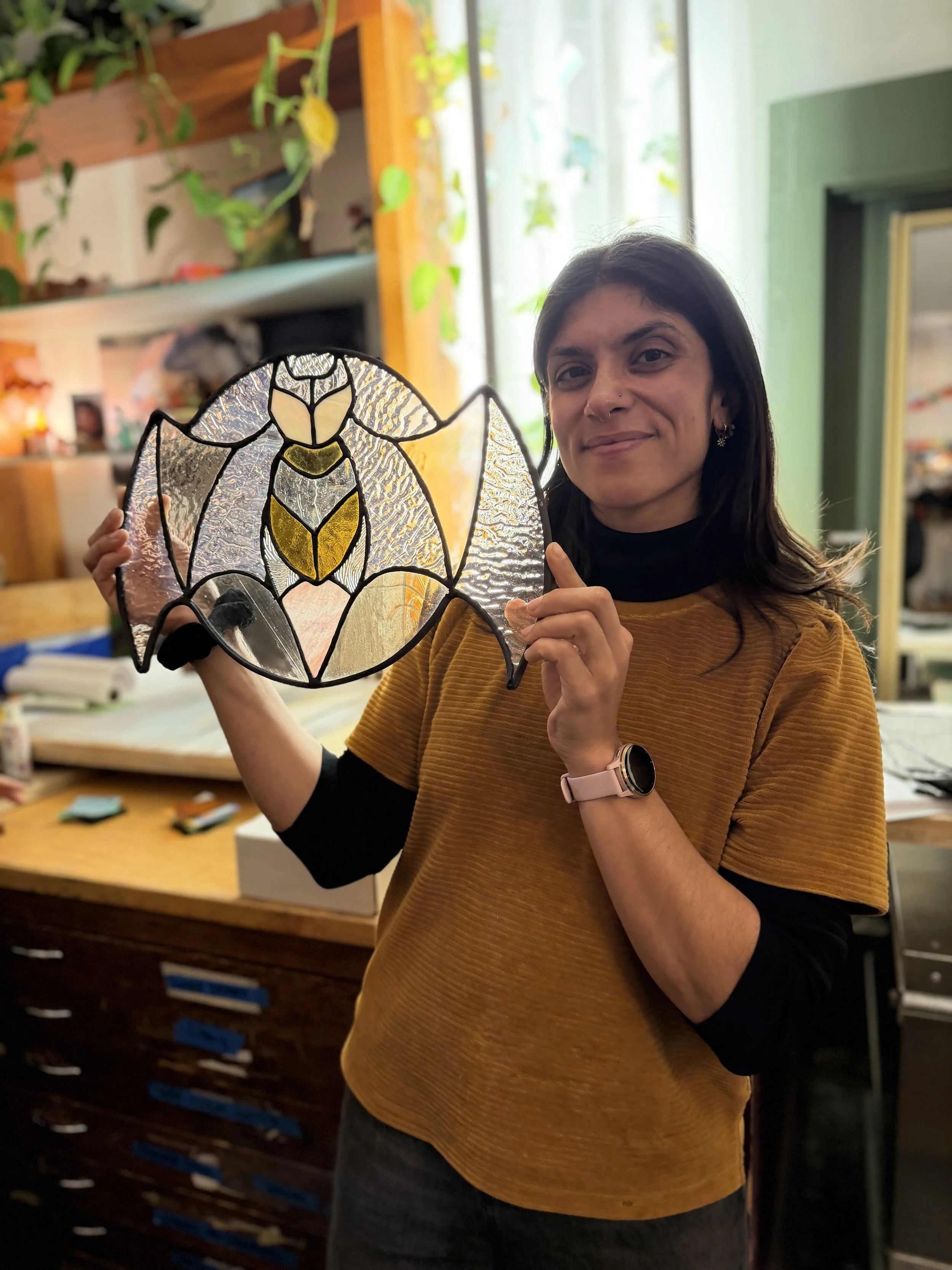 A woman with dark hair holding up a stained glass piece shaped like a butterfly or bee in a craft or art studio.