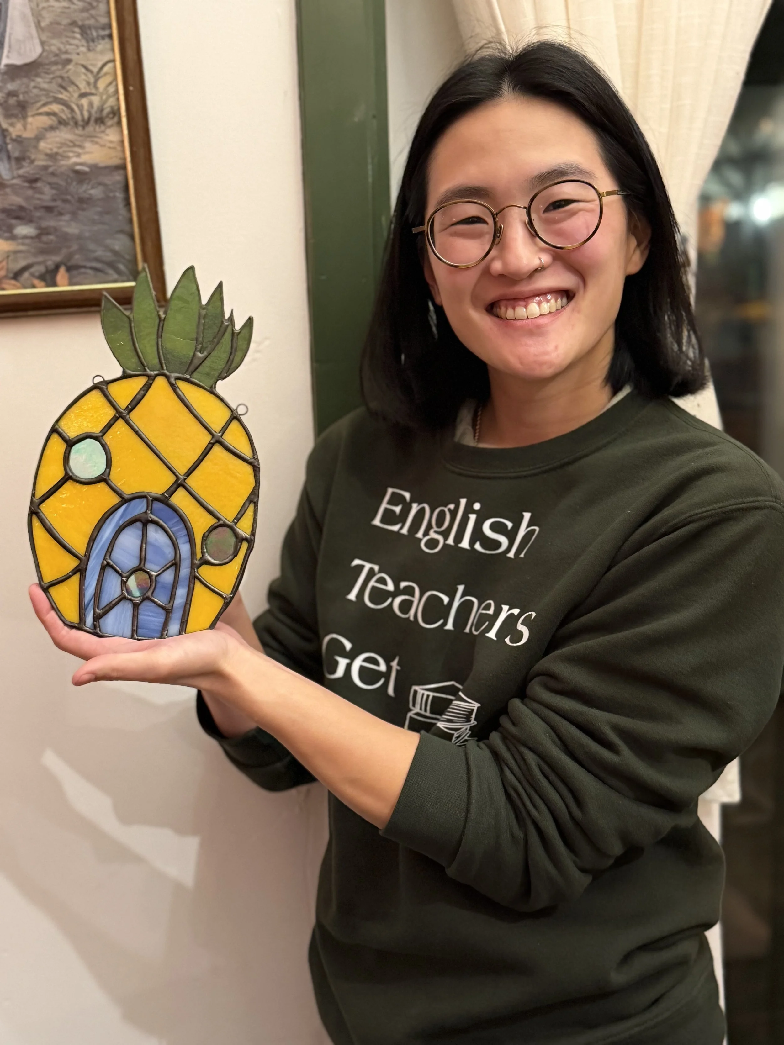 A young woman is smiling and holding a stained glass pineapple decoration.