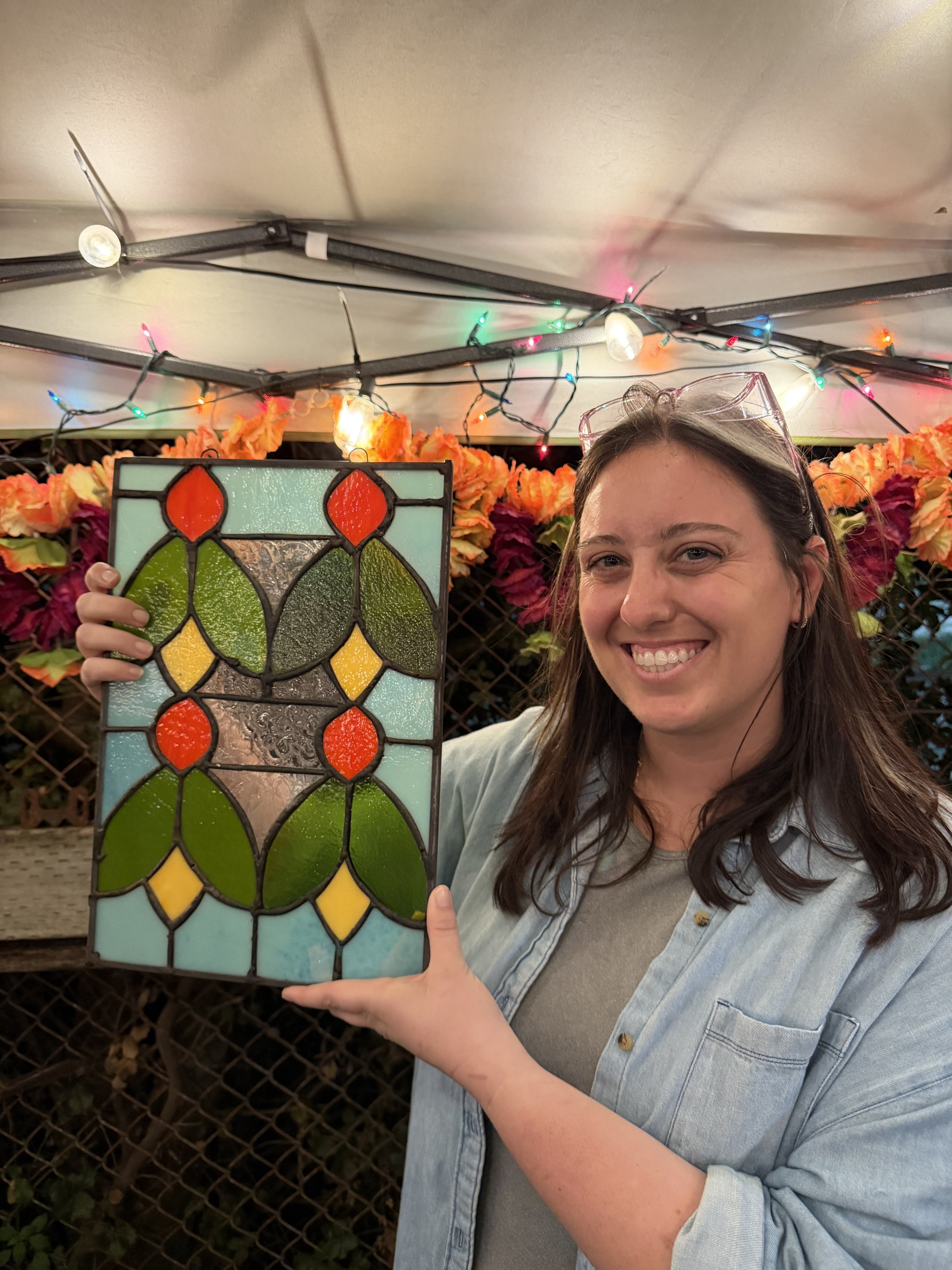 A woman smiling and pointing at a stained glass panel she is holding, with colorful flowers and string lights in the background.