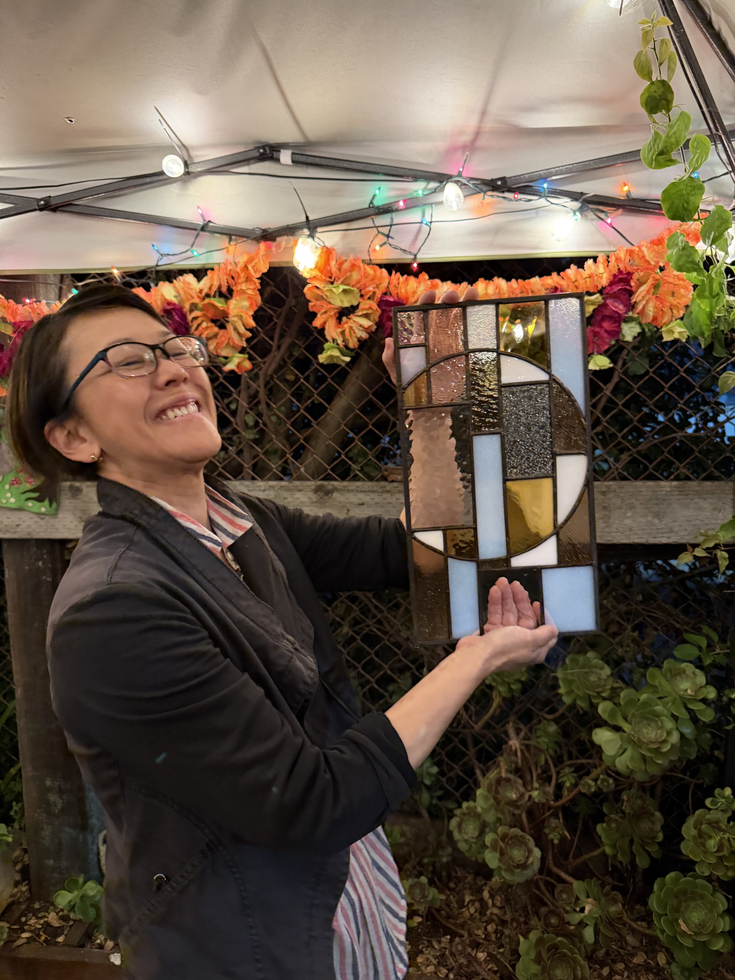 A woman is smiling and holding a stained glass panel in front of a garden with green plants and flowers. There are multicolored string lights and a white canopy above.