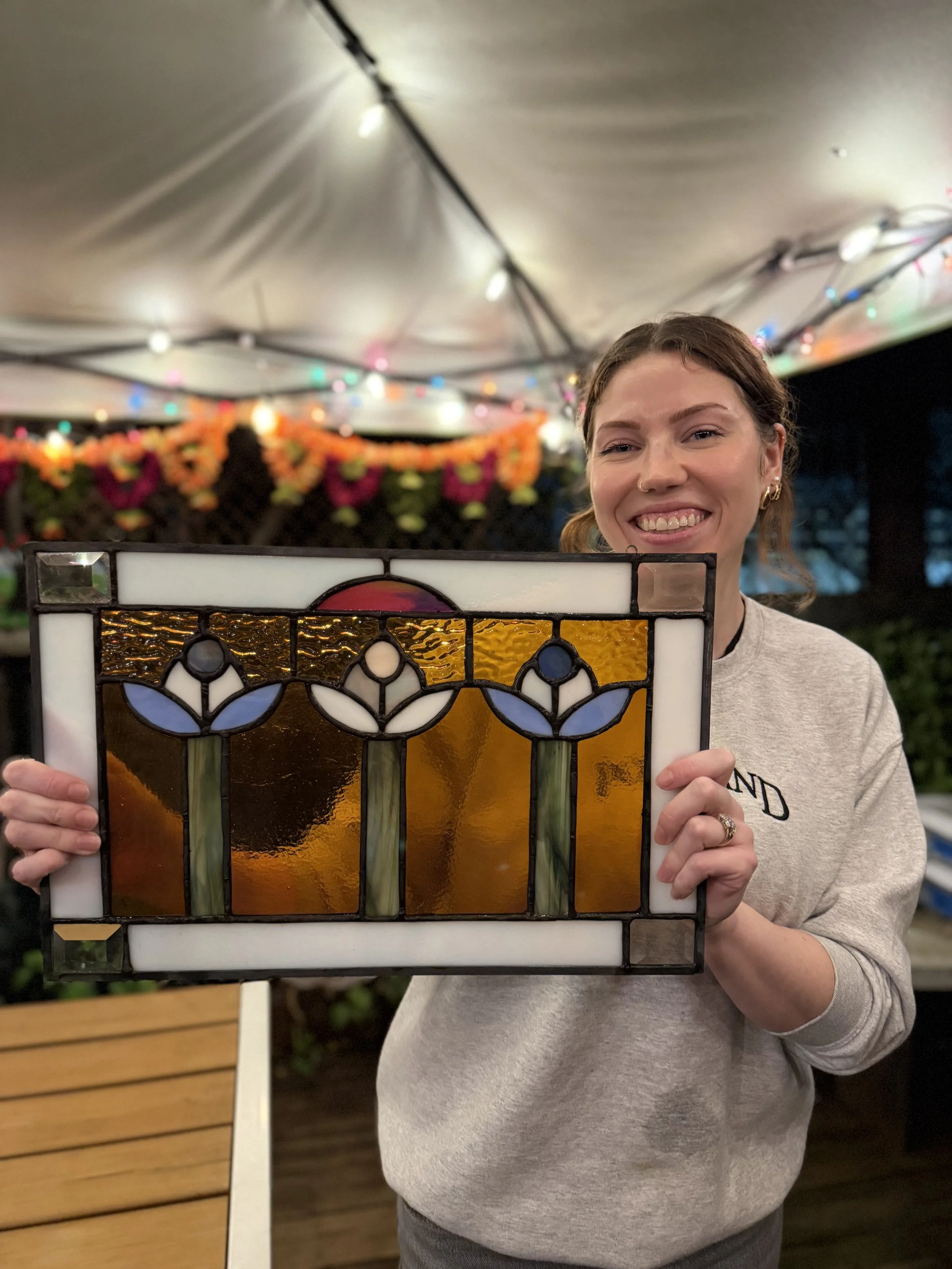 A smiling woman holding a stained glass piece with floral design, in an outdoor setting decorated with string lights and flower garlands.