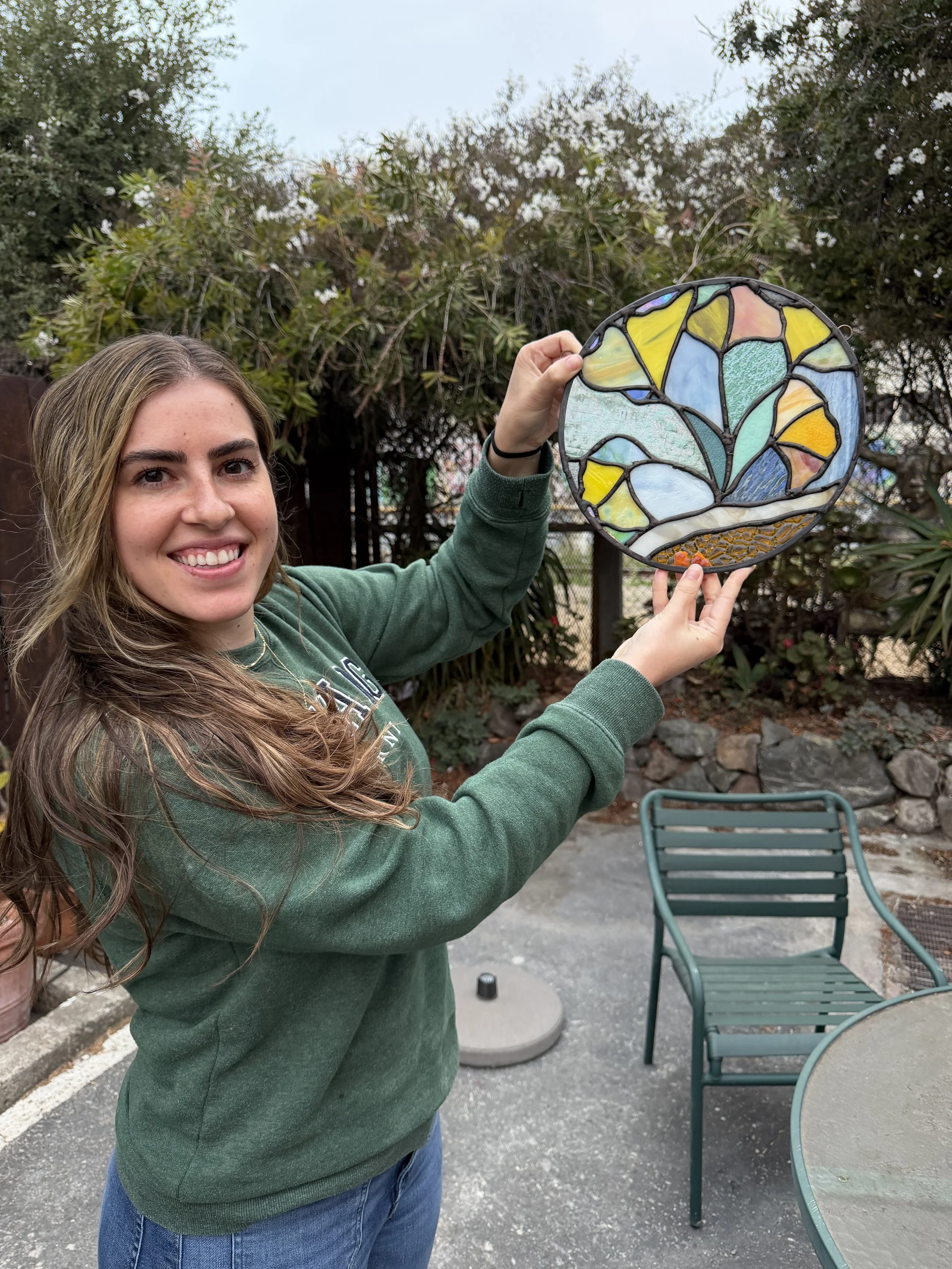 A young woman smiling outdoors, holding up a colorful stained glass circular artwork in front of her. She is dressed in a green sweatshirt and jeans, with a background of trees, a patio, and outdoor furniture.
